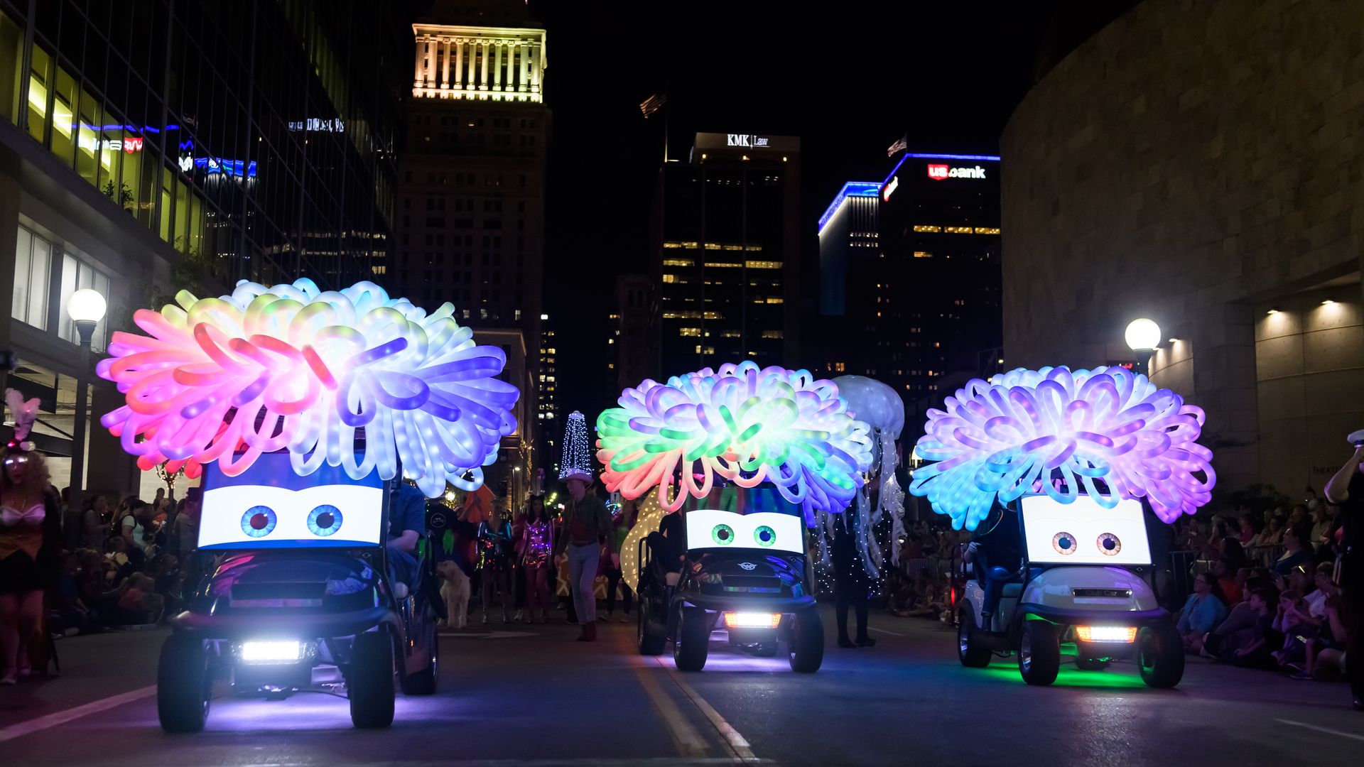 Nighttime city parade featuring glowing rainbow balloon sculptures atop toy cars with cartoon faces, crowds along the street, and illuminated office buildings with a visible PNC sign.
