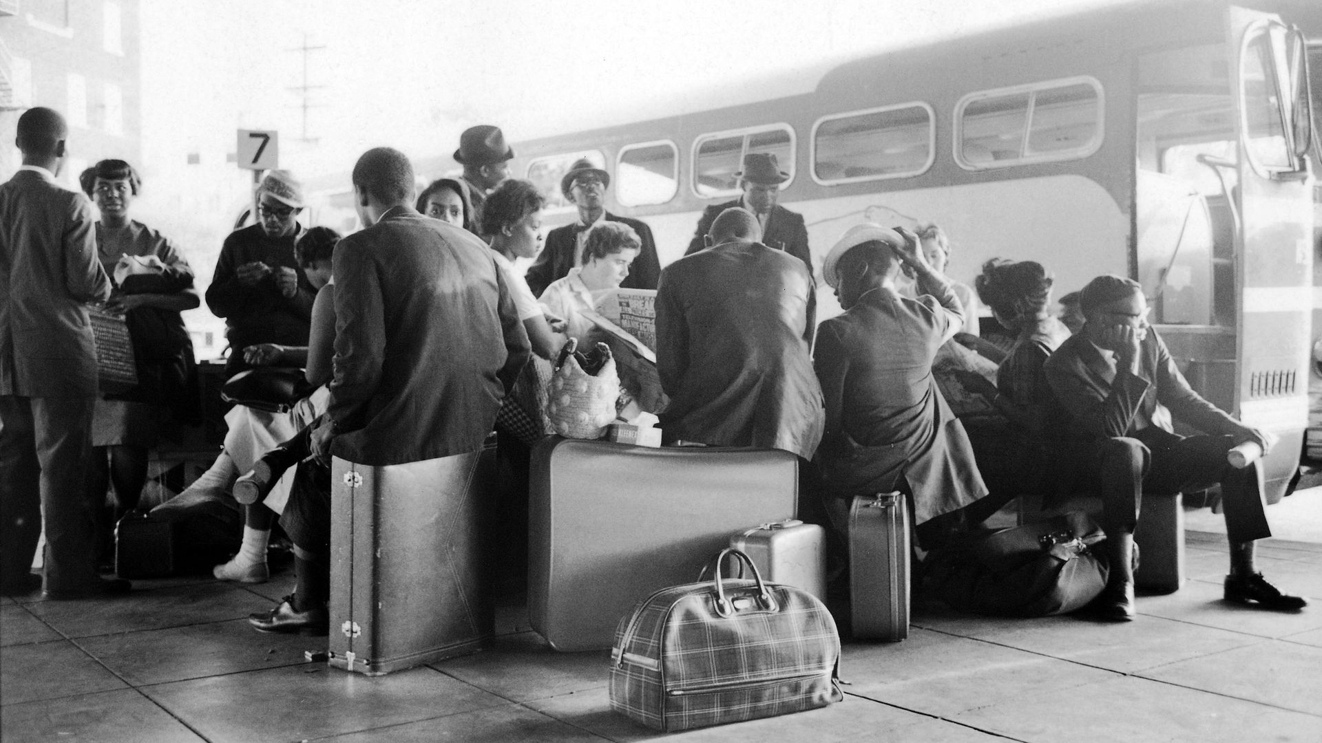 A black and white photo of a diverse group of people standing and sitting atop luggage next to a bus.