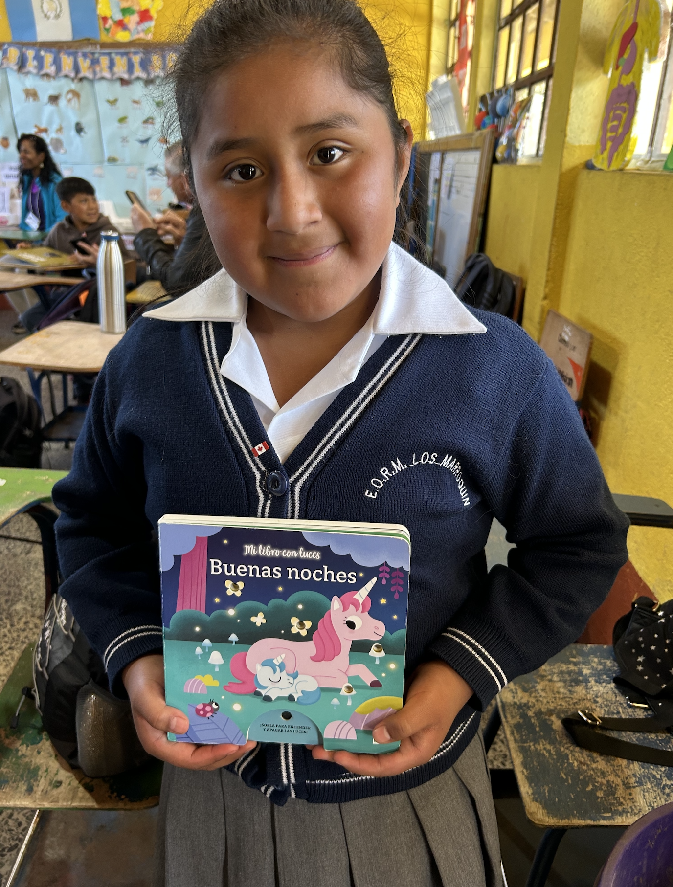 Smiling young girl in a navy cardigan and white collared shirt holds a colorful book titled "Buenas noches" with unicorns on the cover, in a bright classroom with other students and yellow walls.