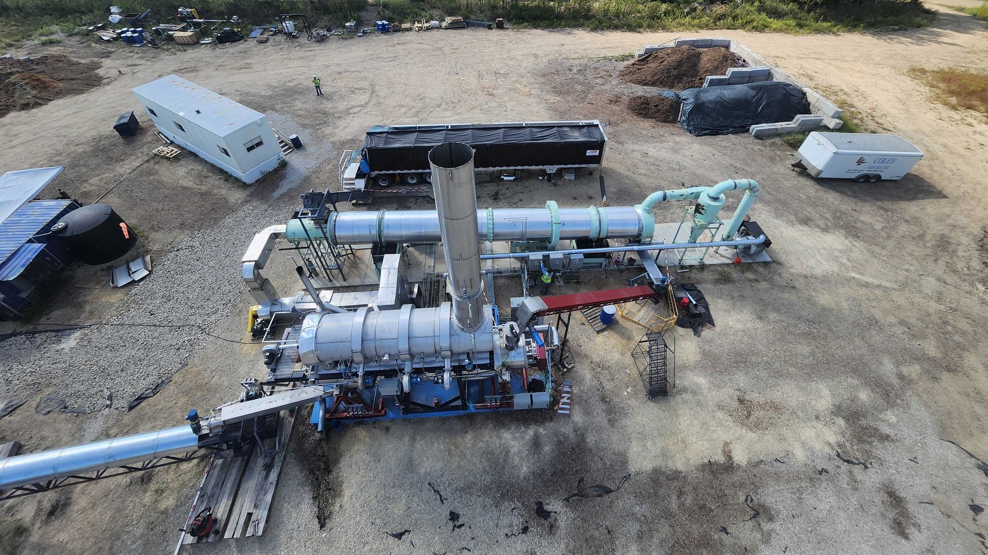 Aerial view of an industrial yard with large piping and machinery, pallets and containers on dirt ground, a worker in a safety vest, and dense trees behind, with distant buildings.