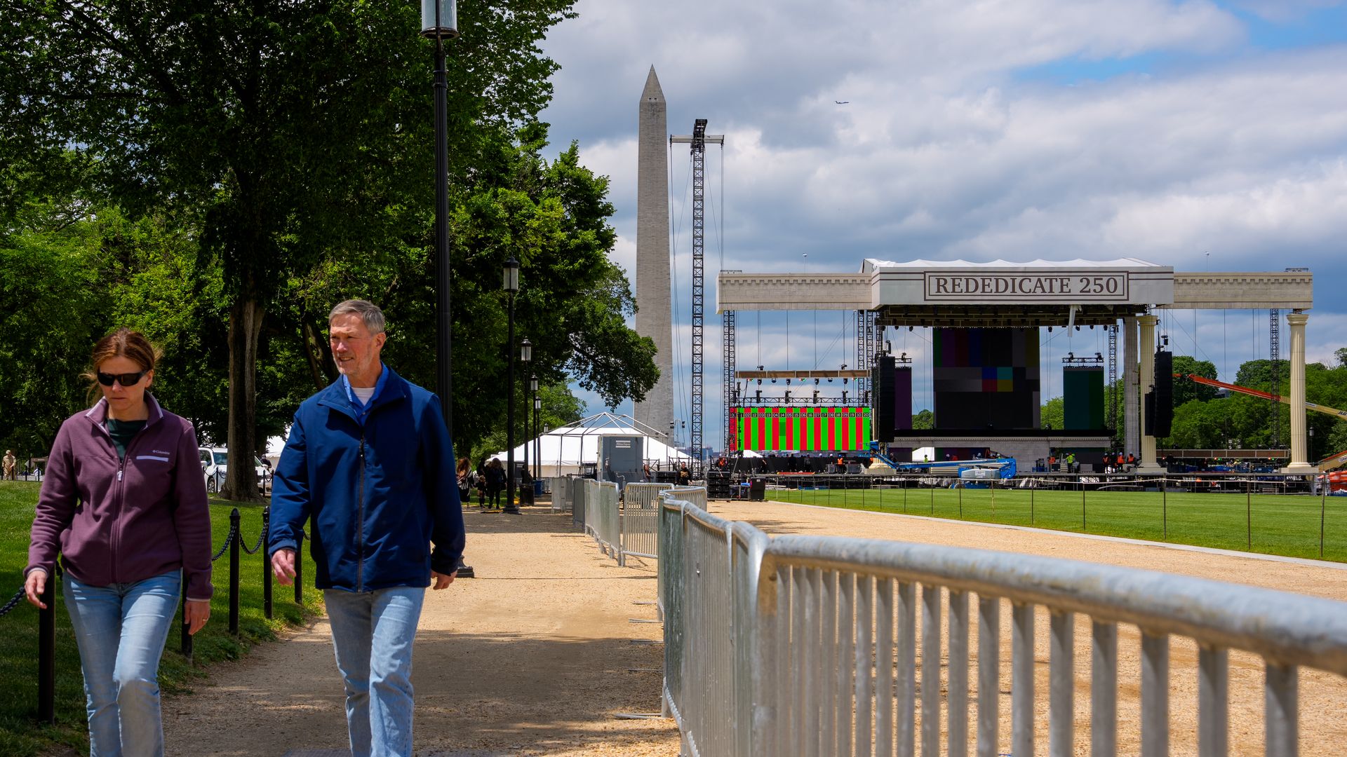 Two people walk along a gravel path beside a barrier toward a large outdoor stage with a banner reading "REDEDICATE 250", a tall obelisk, scaffolding, and green trees on the left under a cloudy sky.