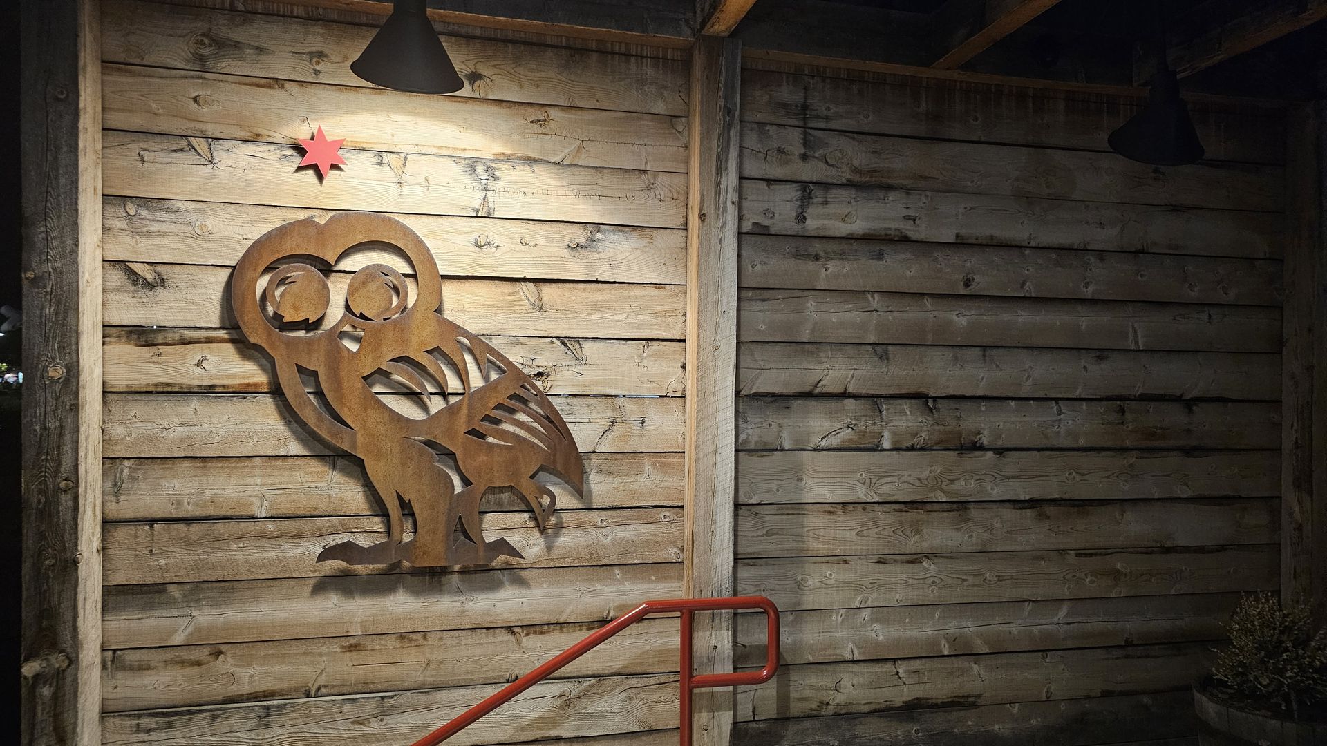 Rust-colored metal owl art mounted on a light wooden wall with a red star above it, illuminated by a black lamp overhead; partial view of a red handrail and wooden barrel with dried plants.