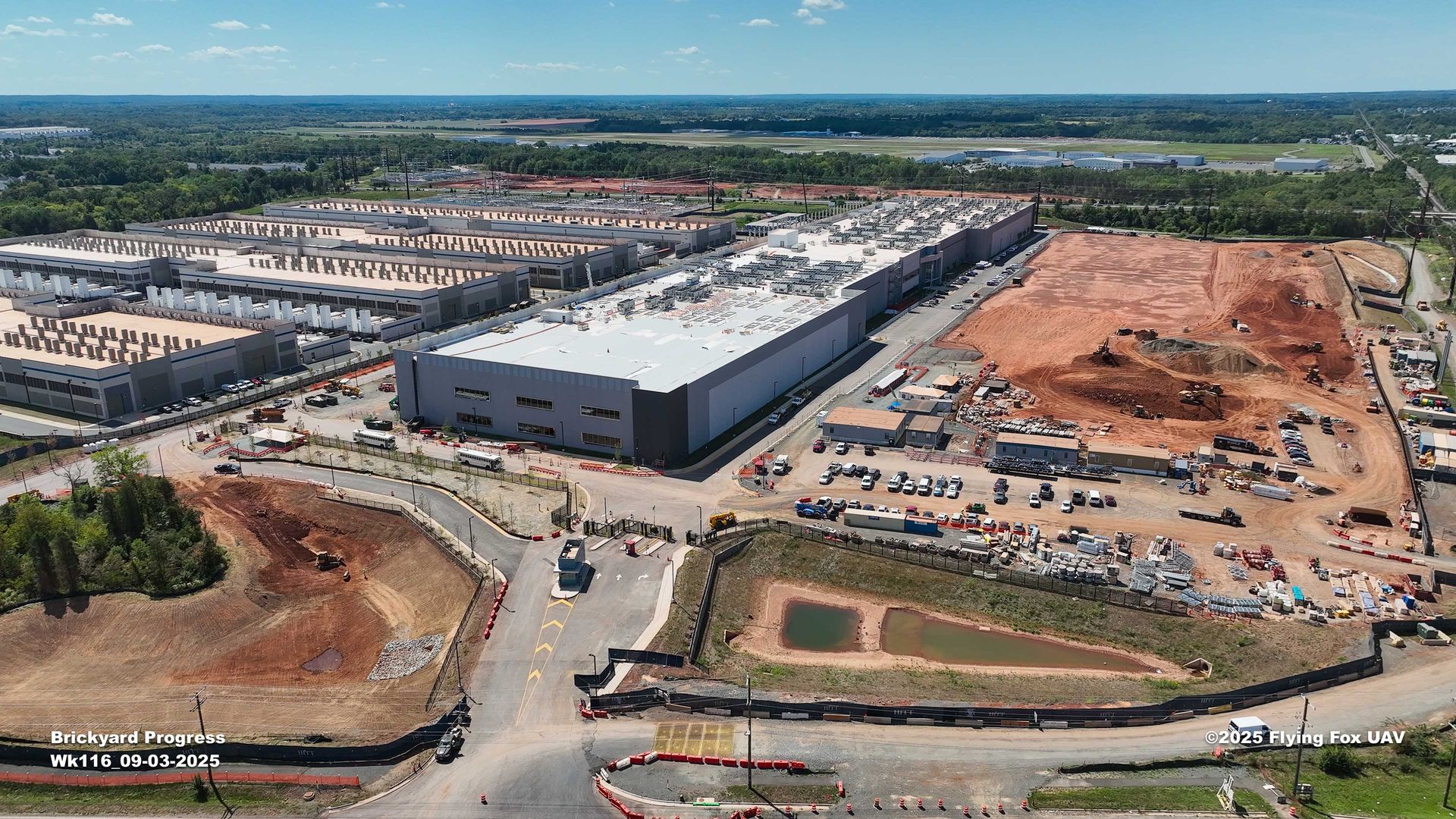 Aerial view of a large industrial construction site with multiple large gray buildings, dirt areas under development, parked vehicles and equipment under a clear blue sky.