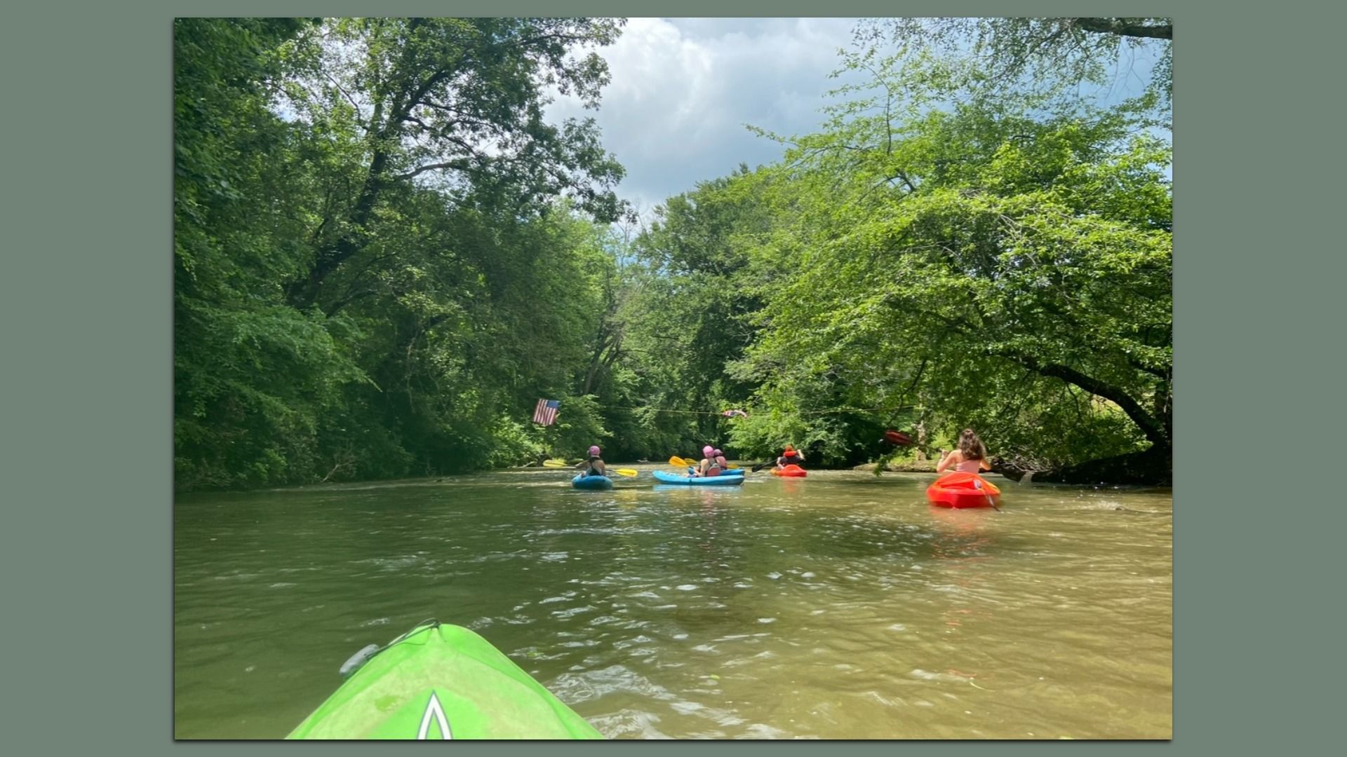 A photograph shot from the perspective of a person in a kayak in a river surrounded by green large trees. Other kayakers are in the distance.