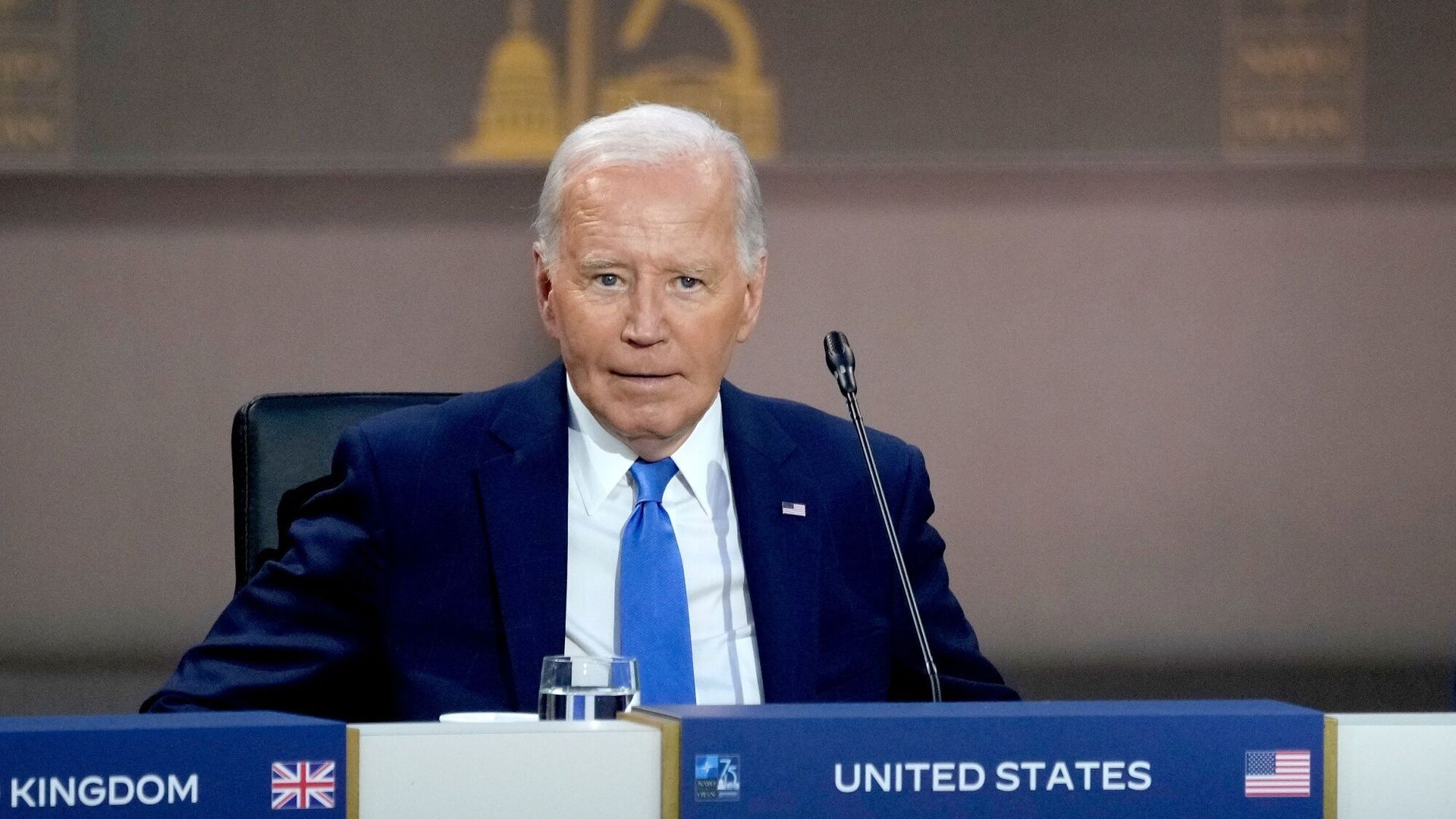 President Joe Biden, wearing a blue suit and sitting in front of a microphone and a "United States" placard.