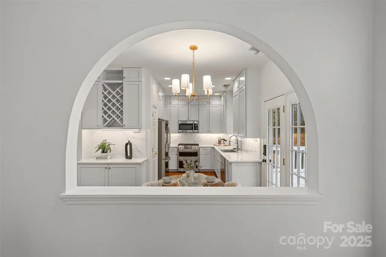 View of a modern white kitchen with gray cabinets, marble countertops, a gold chandelier, stainless steel appliances, and a round dining table set with dishes, seen through an arched wall opening.