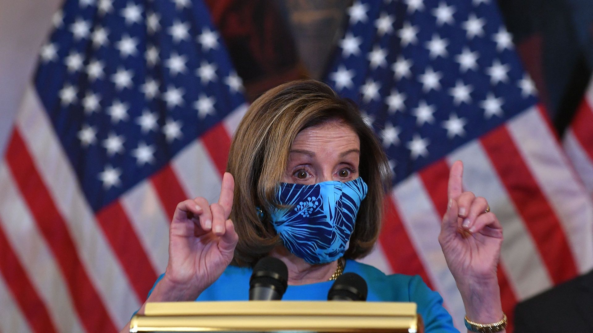 Nancy Pelosi wears a mask while standing in front of American flags