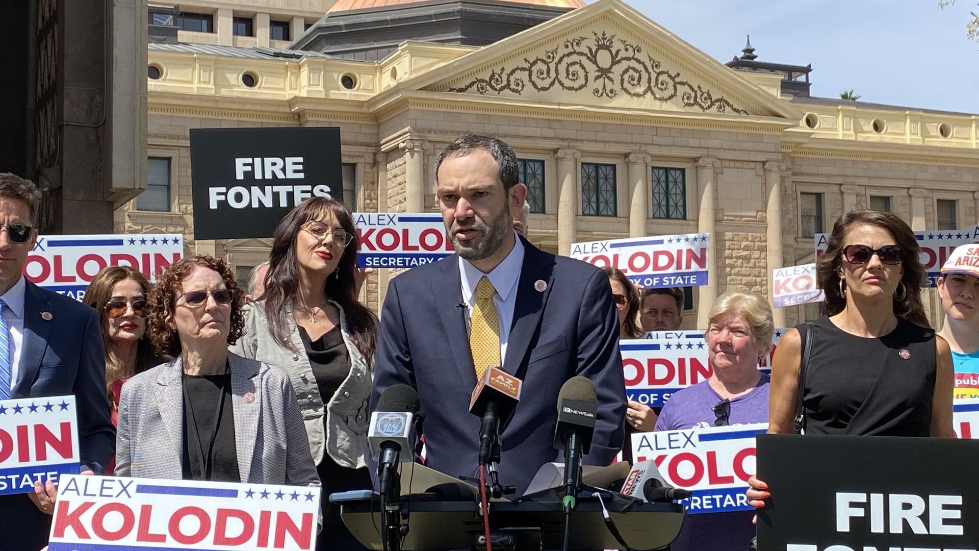 Alex Kolodin speaks into microphones in front of the Arizona state Capitol, surrounded by people waiving signs that read "Alex Kolodin Secretary of State" and "Fire Fontes".