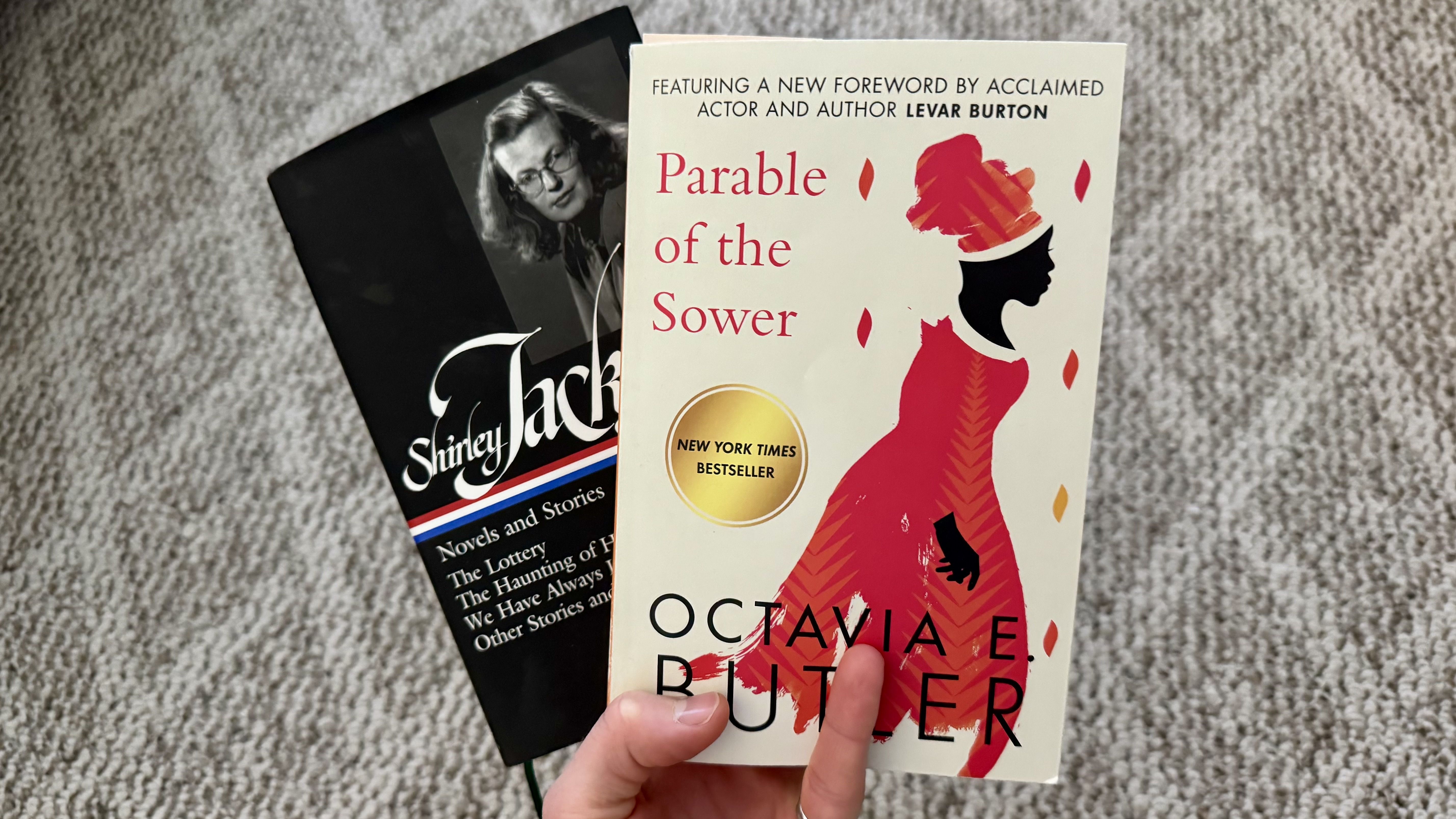 Hand holding the book "Parable of the Sower" by Octavia Butler with a white cover and red silhouette of a woman, placed over a book by Shirley Jackson.