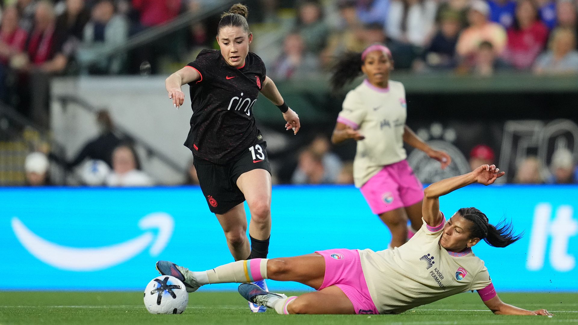 Women’s soccer action: a black-kit player (number 13) dribbles the ball as a cream-and-pink defender slides in for a tackle on a green field, with a blurred crowd in the background.