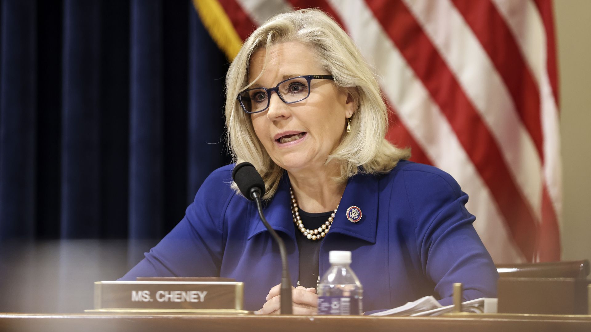 Rep. Liz Cheney (R-Wyo.) in a blue blazer sits at a table, behind a microphone and a placard of her name, and in front of an American flag.