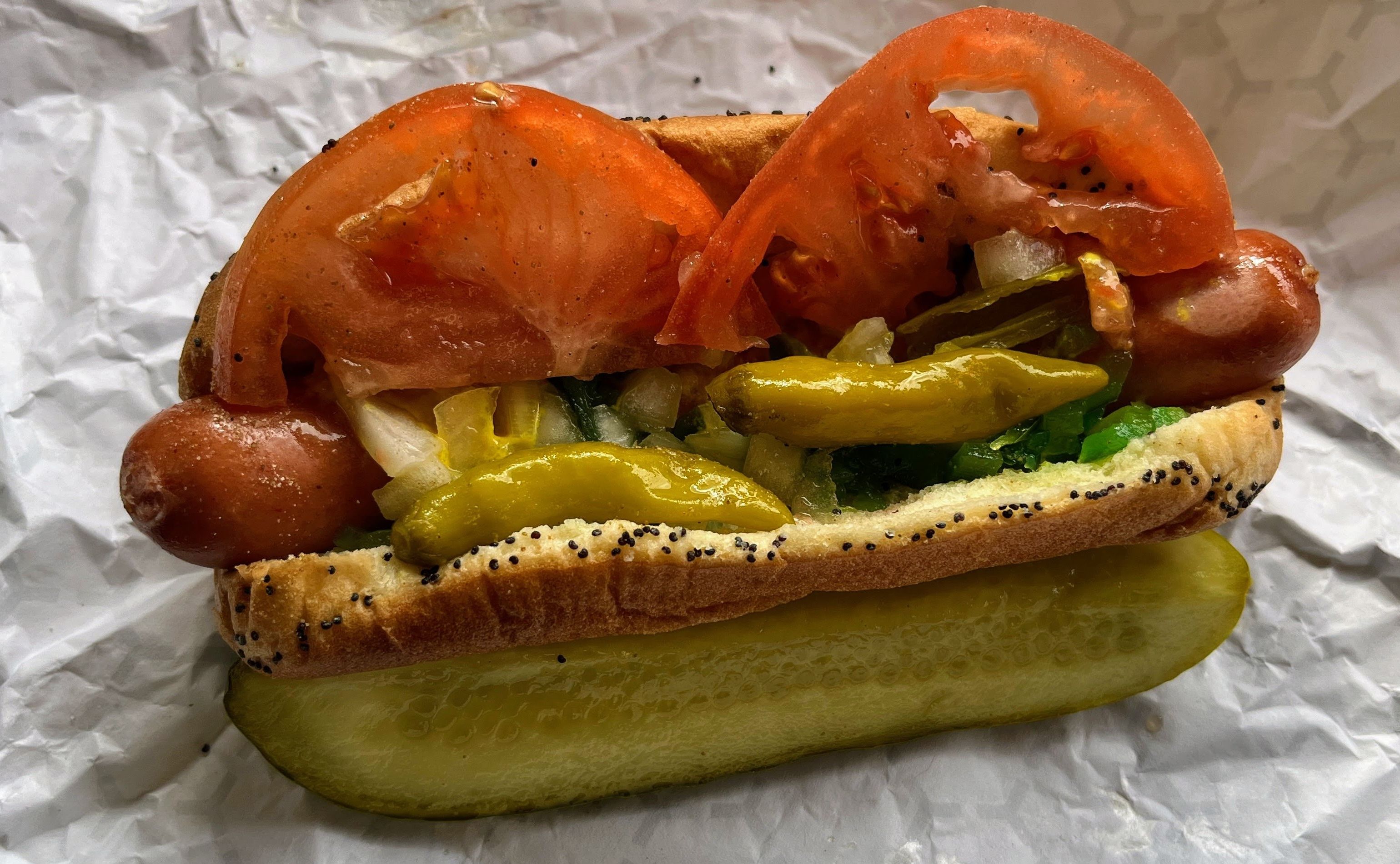 Close-up of a hot dog in a poppy-seed bun, loaded with pickles, orange tomato slices, onions, yellow peppers, and greens, resting on crinkled foil.