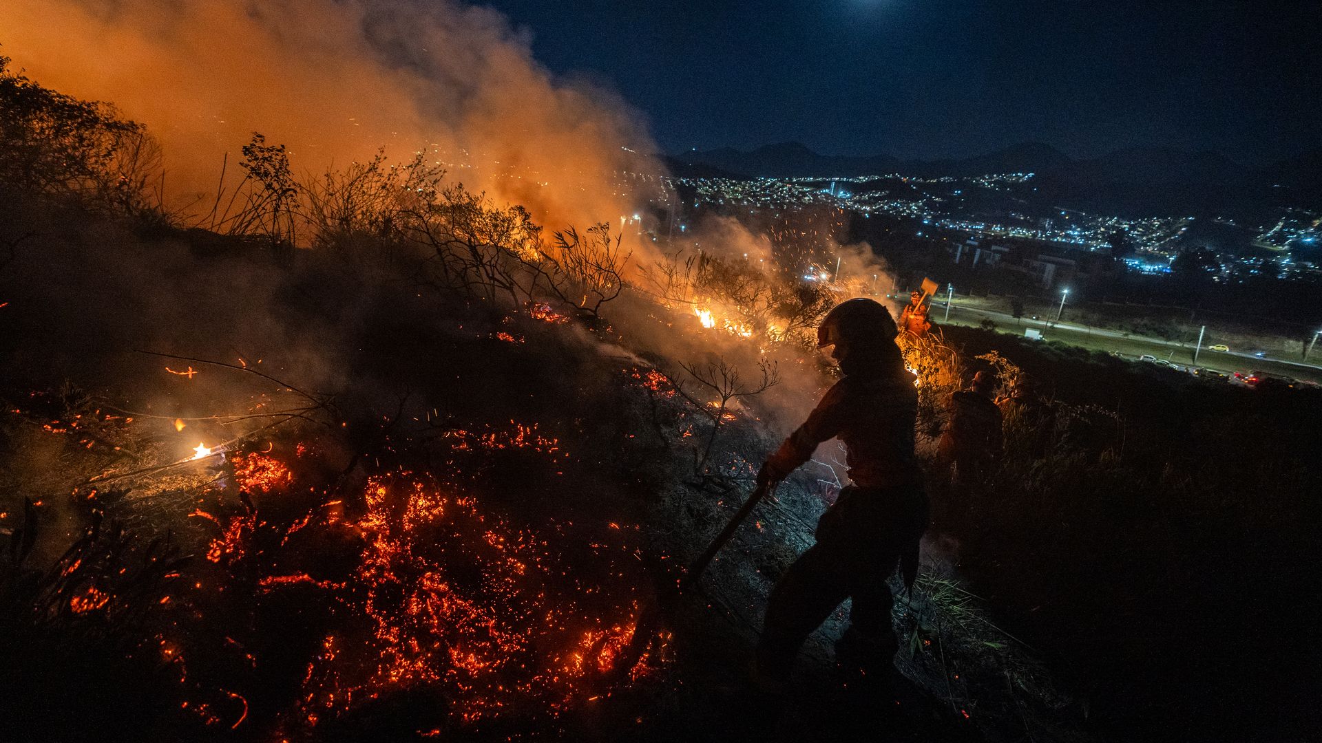 A rescuer working to extinguish a forest fire at night with the lights of Bogota in the background. 