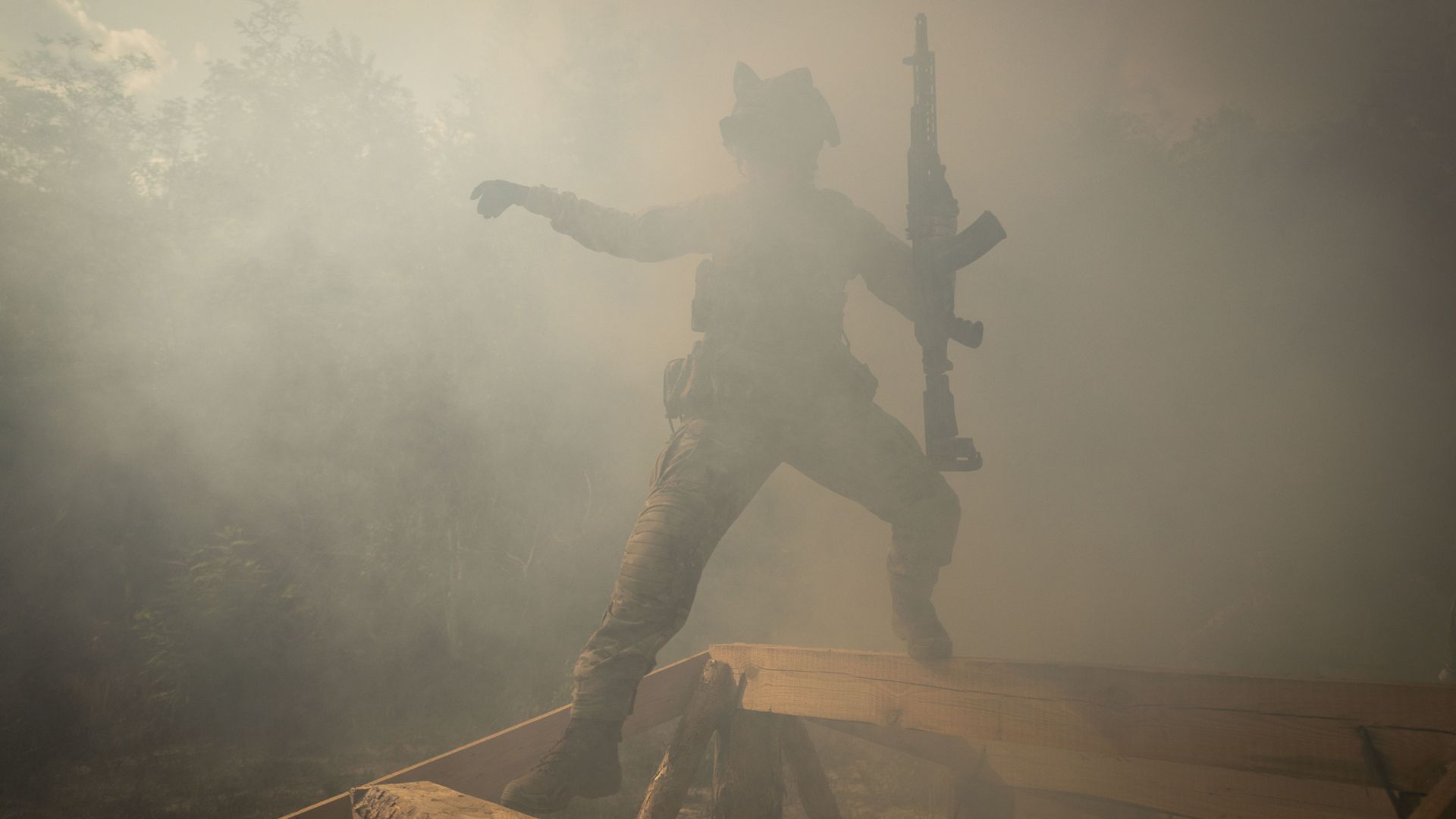 A Ukrainian soldier leaps over an obstacle obscured by smoke. The person is holding a rifle in their left hand.