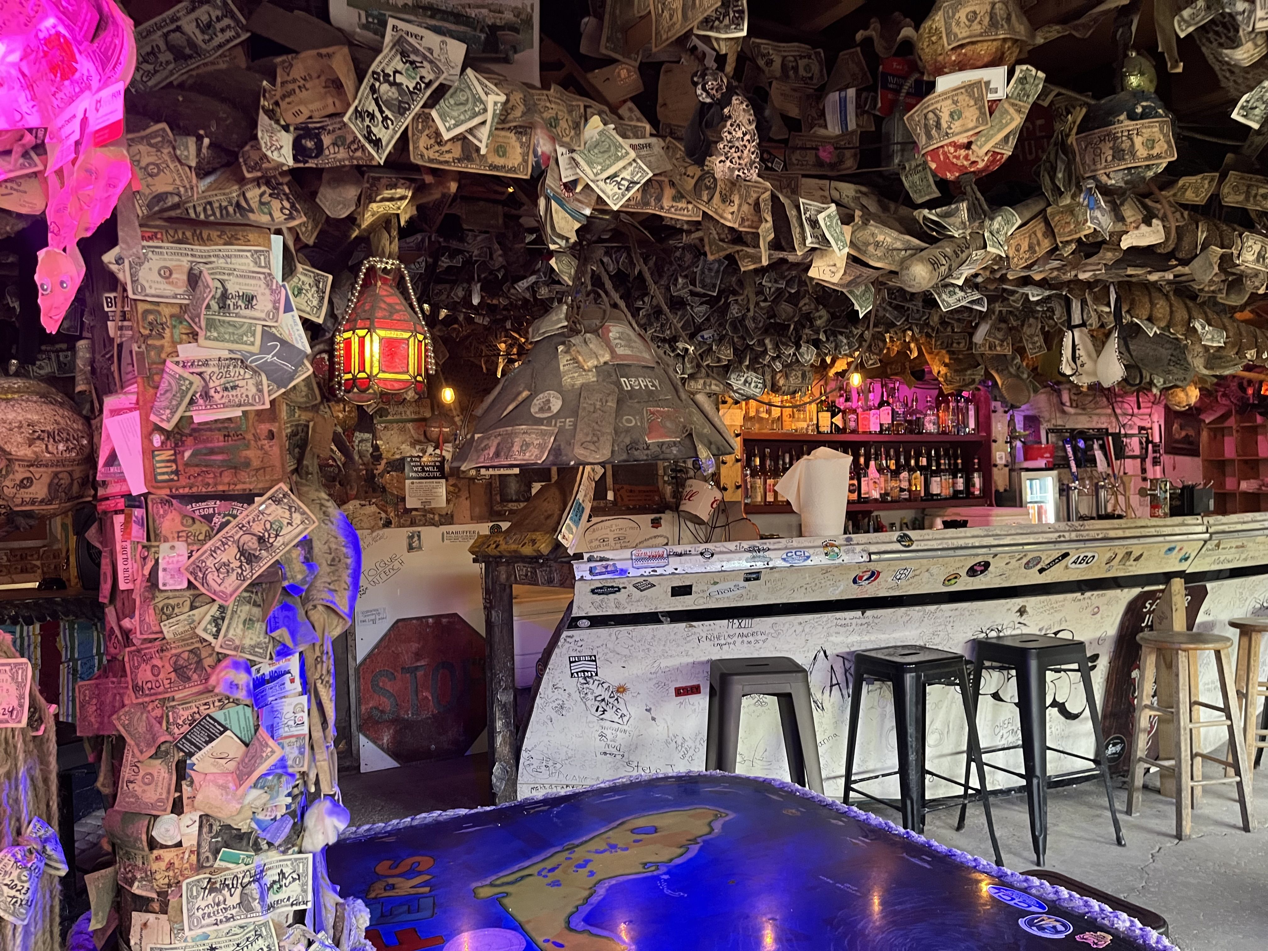 Interior of a quirky bar with dollar bills and notes covering walls and ceiling, colorful hanging lamps, bar stools, and a counter with various liquor bottles in the background.