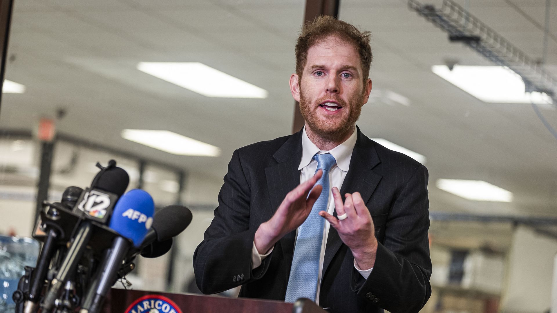 A man in a suit gestures with his hands and speaks in front of several microphones. 