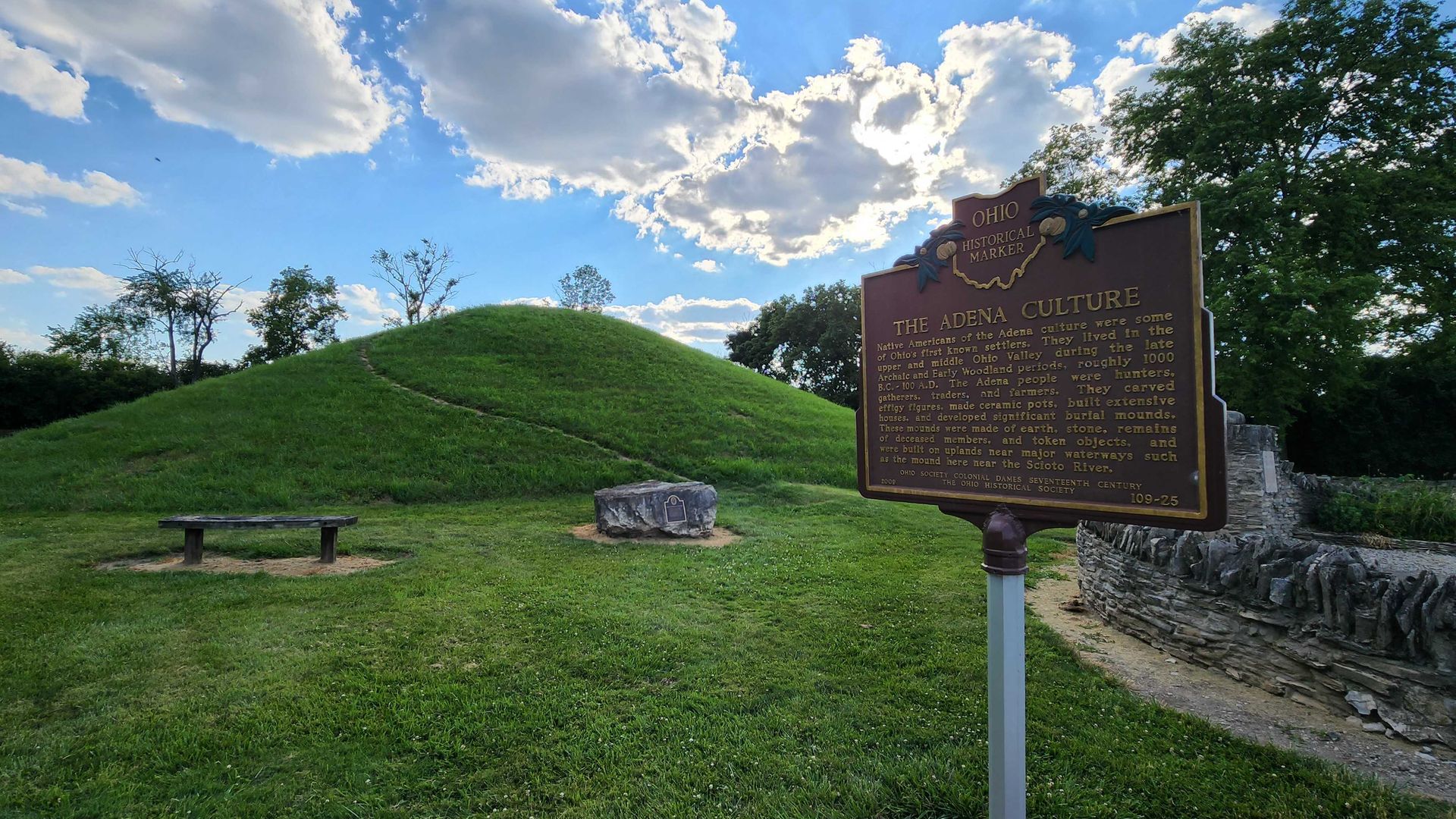 A historical marker sign in the foreground with a burial mound in the background