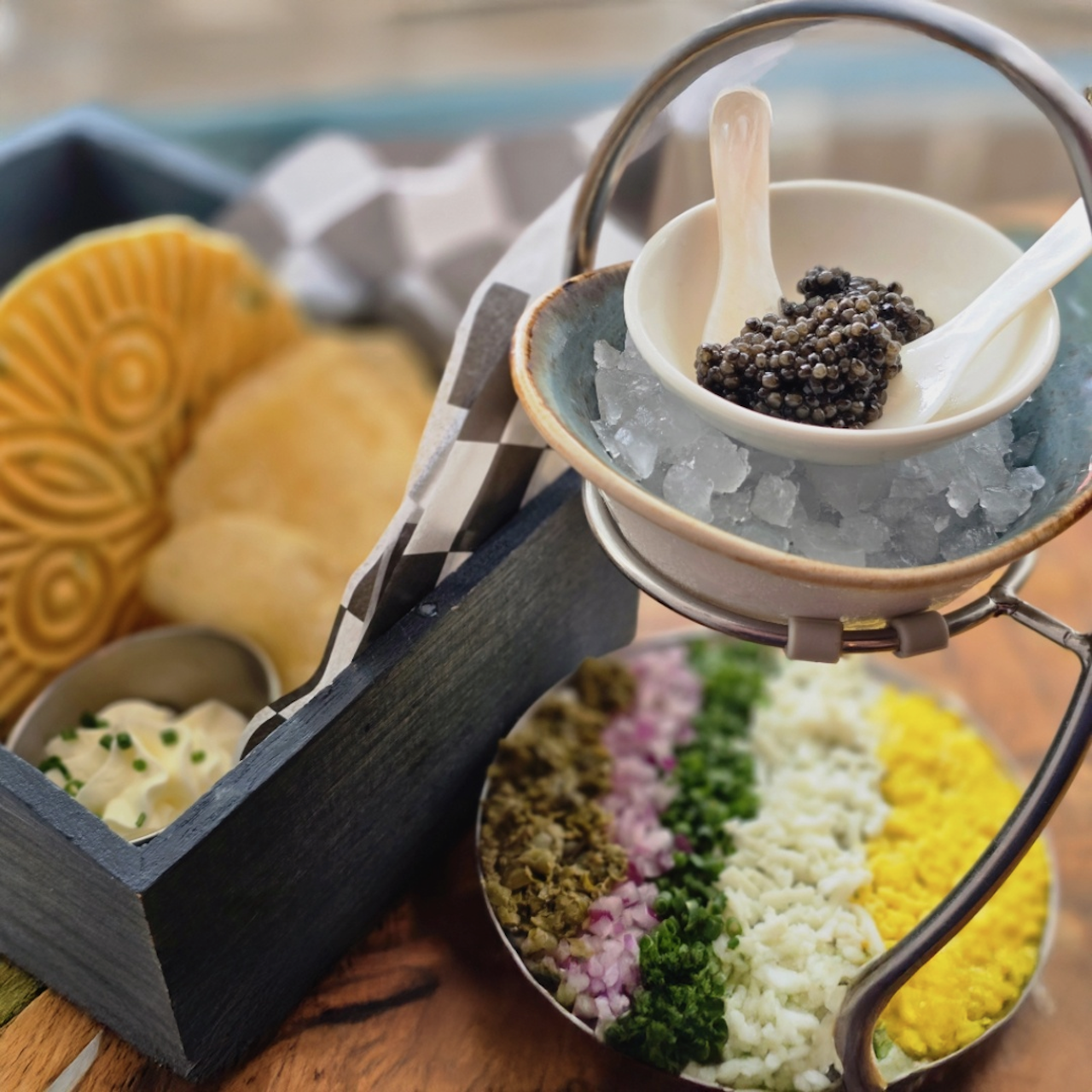 Elegant close-up of black caviar in a white cup on ice, with a white spoon, perched on a metal stand. In the background is a bread basket and a tray of colorful chopped garnish.