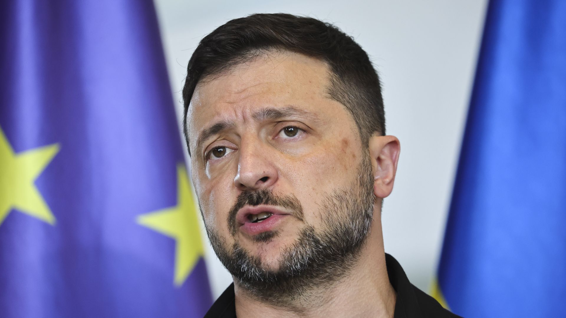 A close-up of Volodymyr Zelensky speaking in front of two blue flags. 