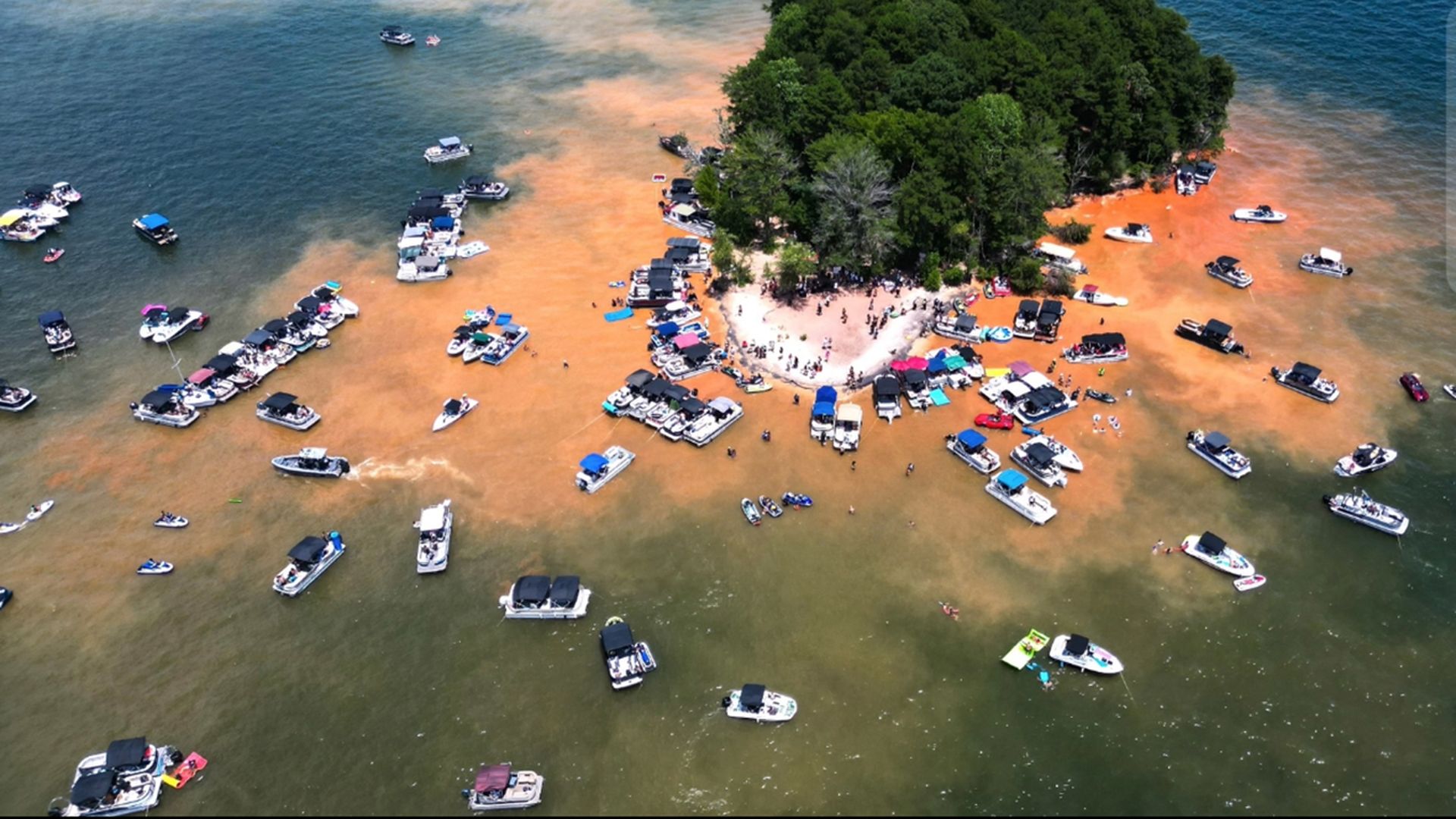 Aerial view of a small green island with a white sandy beach, surrounded by orange-tinted shallow water and dozens of boats anchored around, with a crowd on the shore.