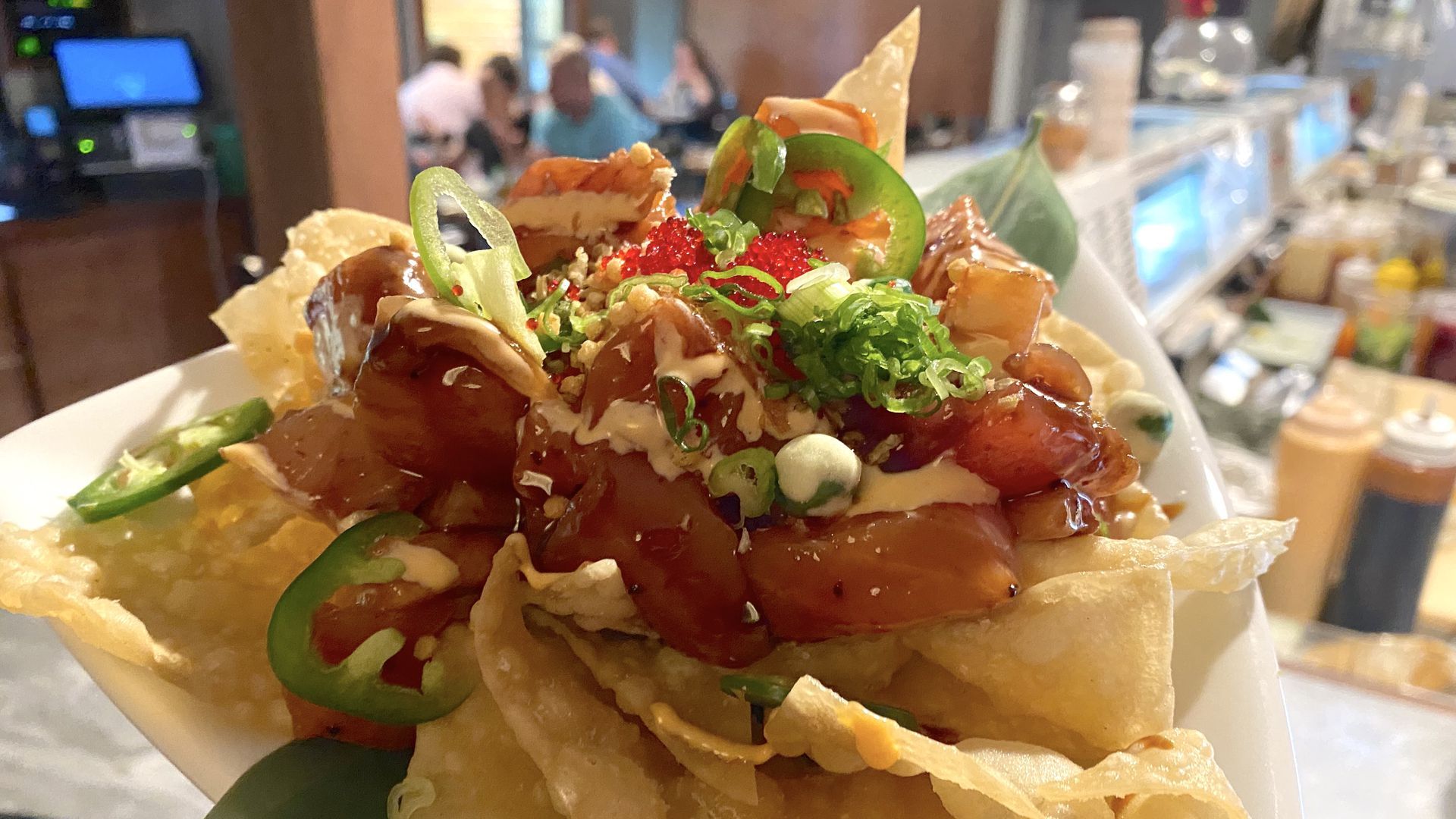A close-up photo of wonton chips covered in sushi, jalapenos, scallions, and sauce.