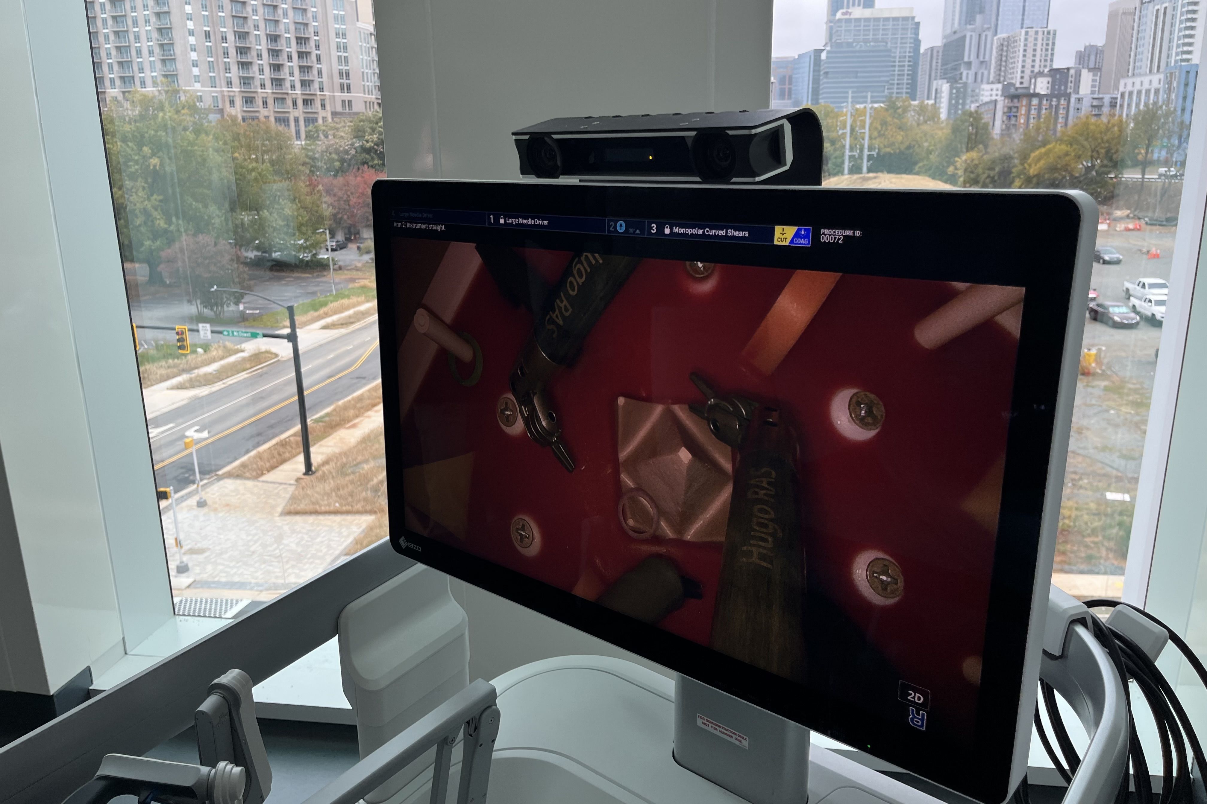 Medical monitor displaying robotic surgical tools with text "Hugo RAS", set near a large window overlooking a city street and buildings on a cloudy day.