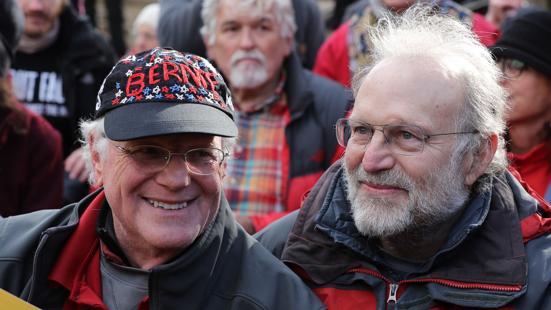 Two older men wearing glasses, one of whom is wearing a hat that reads "BERNIE," smile in a crowd of people.