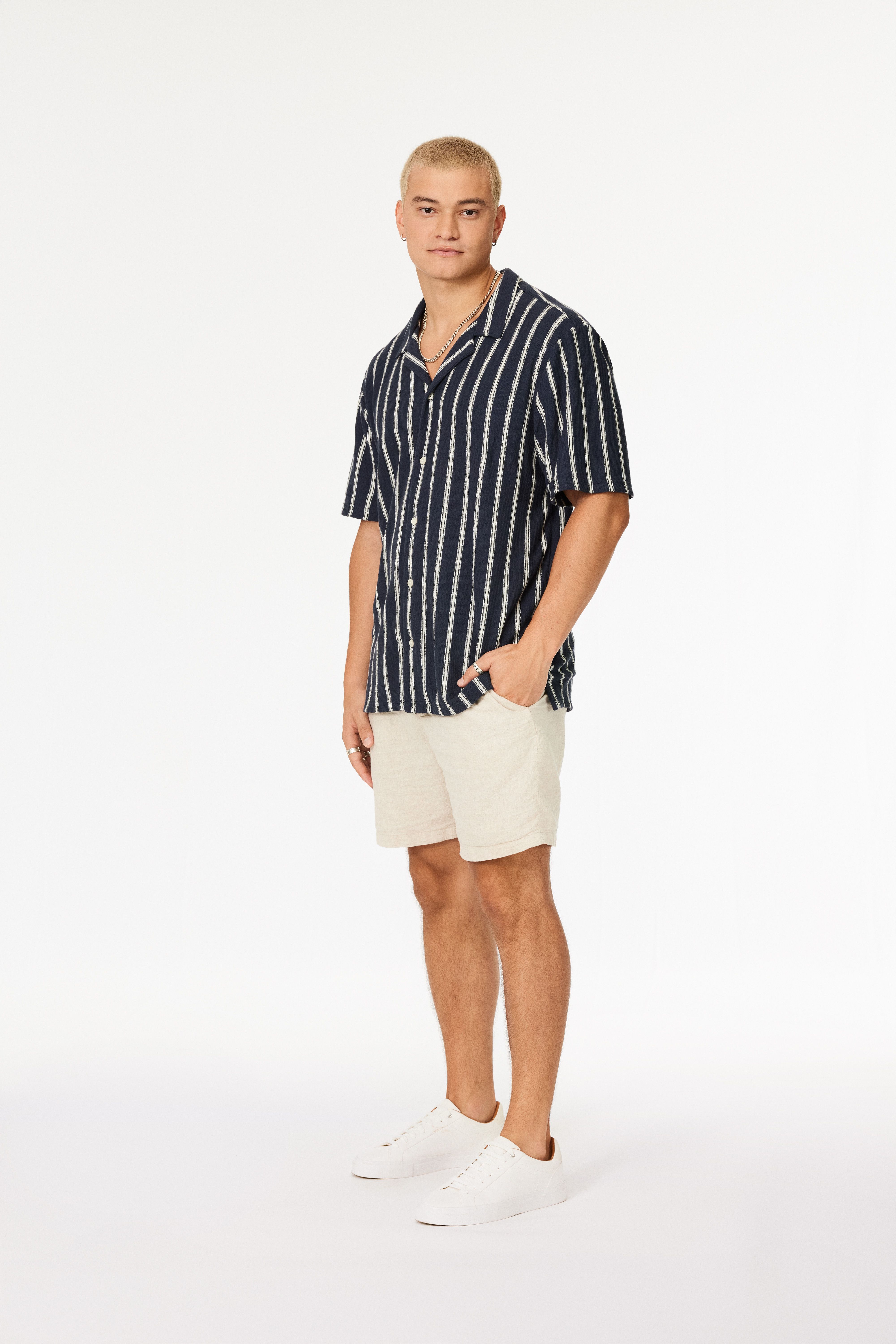 Young man with short blonde hair wearing a navy blue short-sleeve shirt with white vertical stripes, beige shorts, white sneakers, silver chain necklace, and rings, standing against white background.