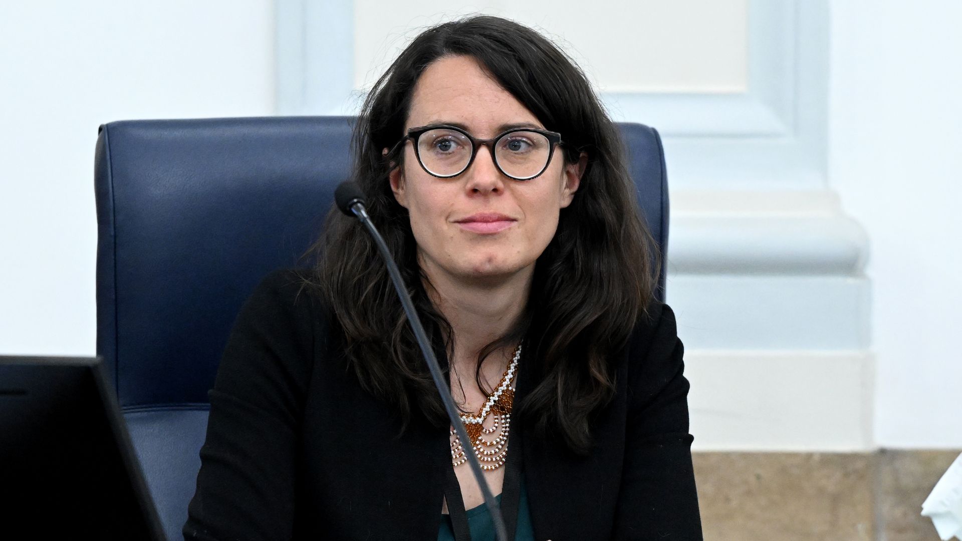 Woman with dark hair and round glasses seated at a judge's bench with a microphone, wearing a black blazer and layered necklace. A nameplate reads PARADY at the front.