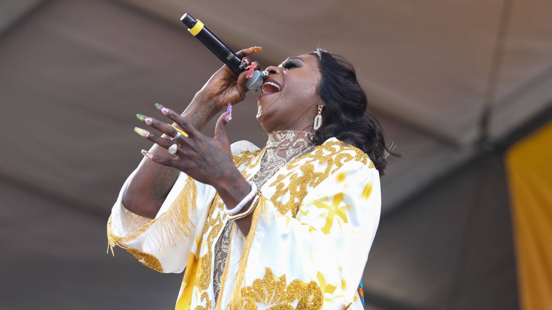 Big Freedia performing with a microphone, wearing a white and gold ornate garment, colorful long nails, and earrings under a tented stage.