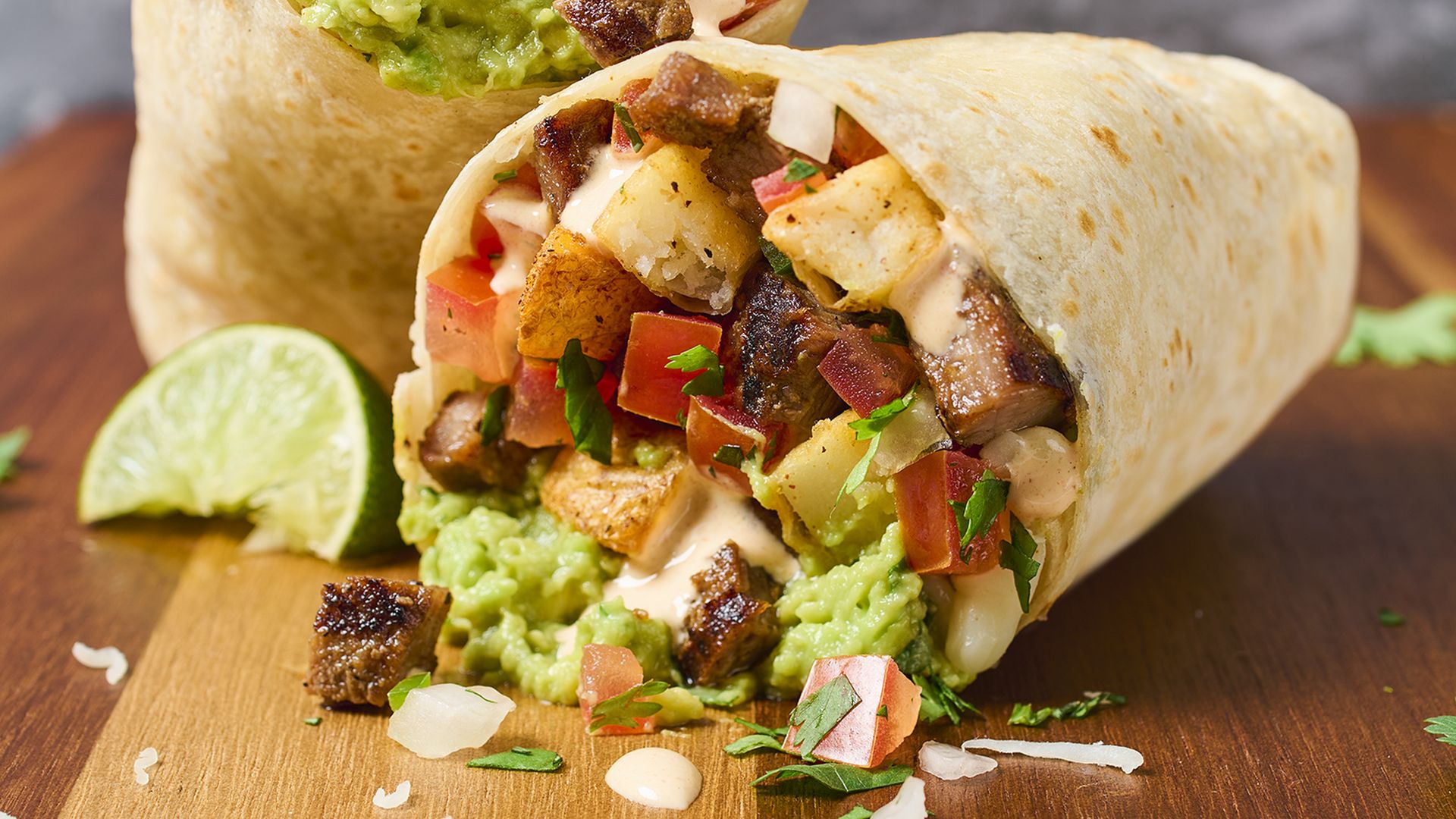 Two halves of a burrito filled with grilled steak, fries, tomatoes, guacamole, cilantro, and cheese, on a wooden surface with a lime wedge and scattered herbs.