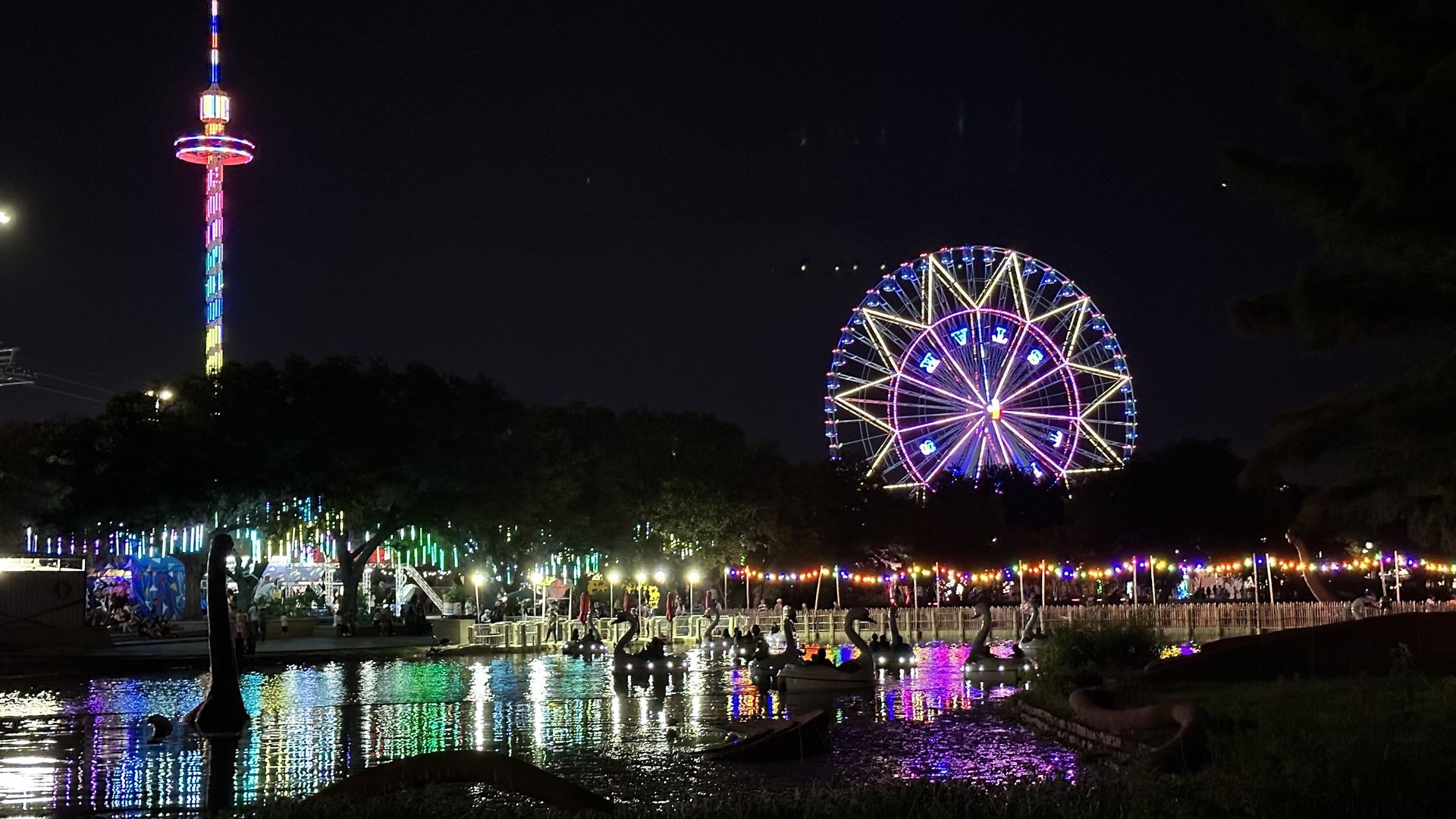 A nighttime photo of a ferris wheel lit up with a pond of ducks in front of it