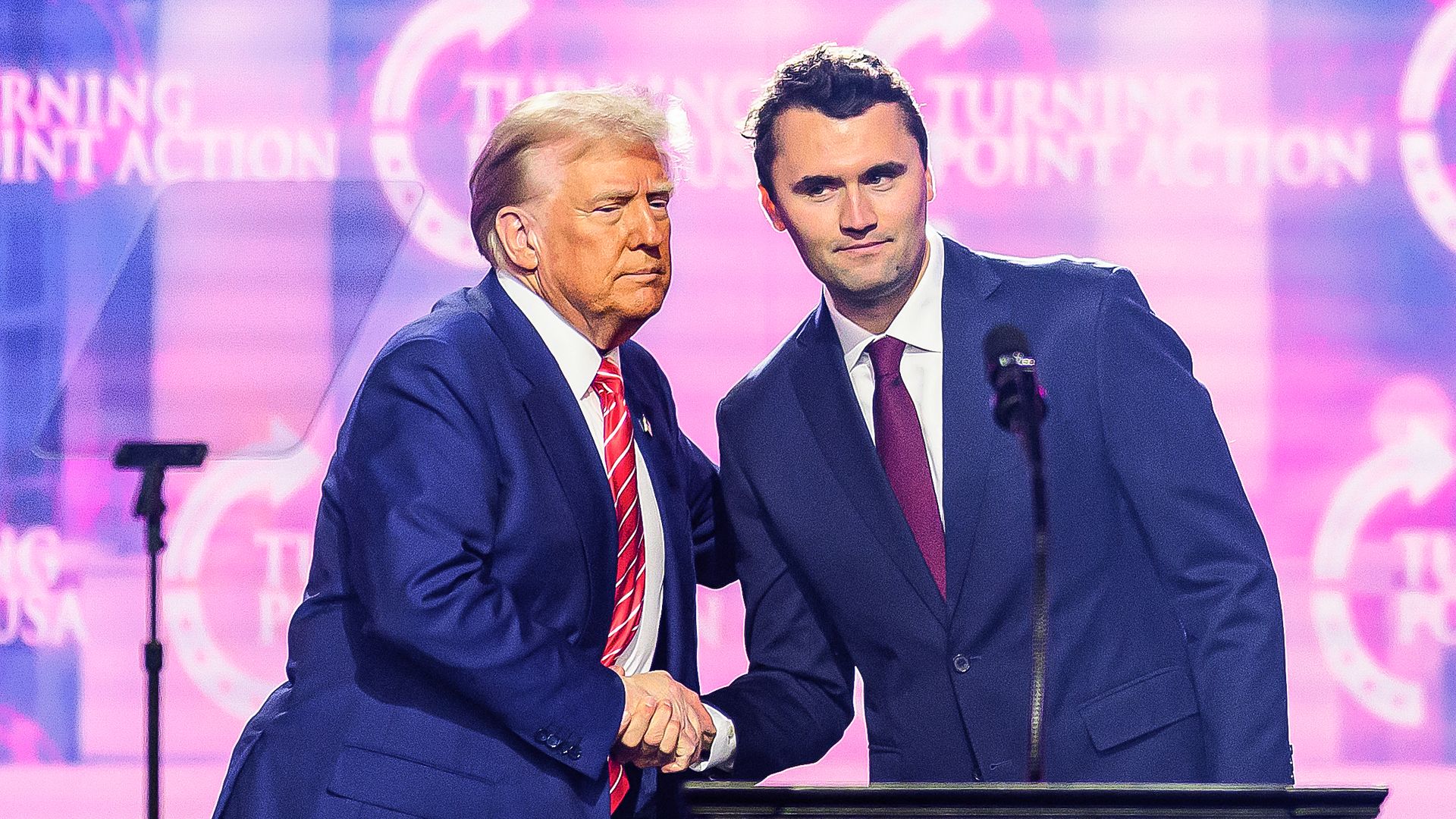 Trump shakes hands with Charlie Kirk behind a podium while a screen behind them reads "TURNING POINT ACTION."