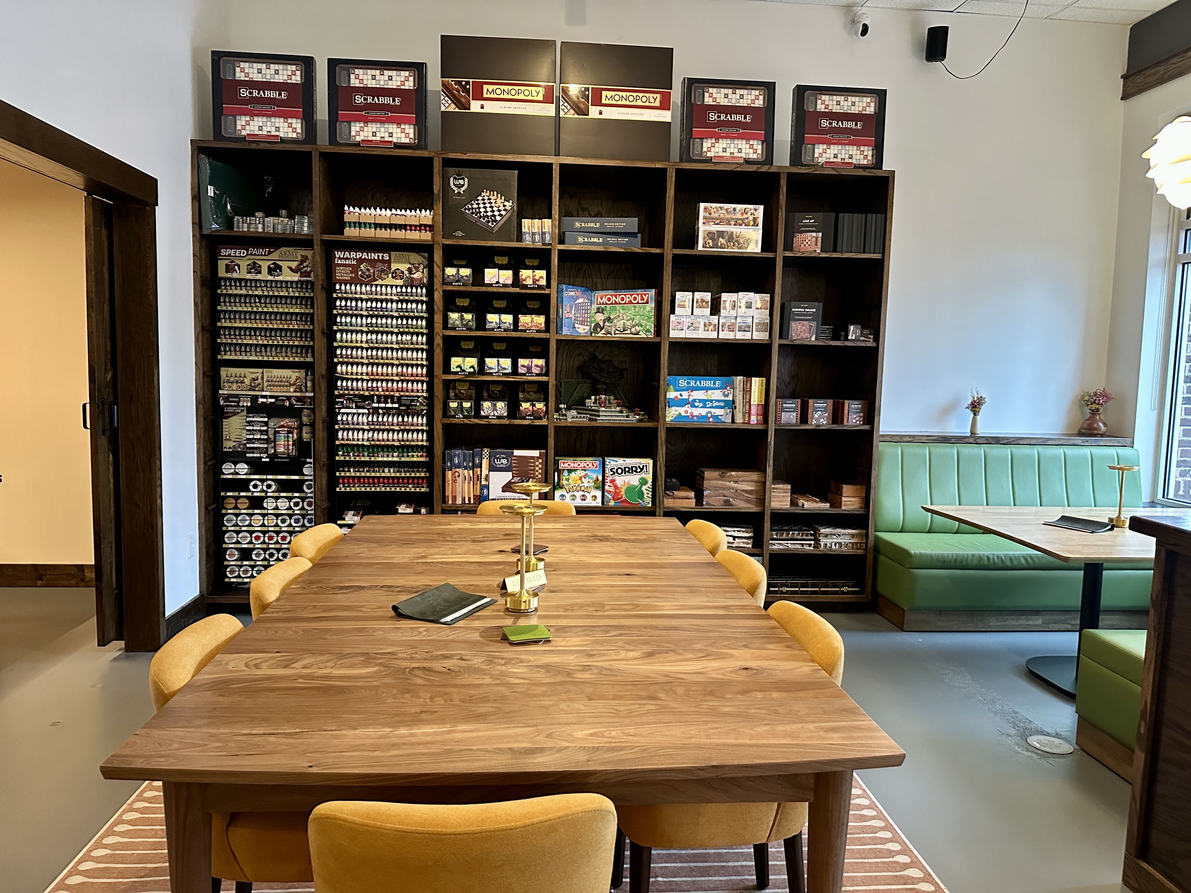 Room with wooden table surrounded by yellow chairs, a green booth, and shelves filled with board games like Scrabble and Monopoly, along with paint supplies and miniatures.