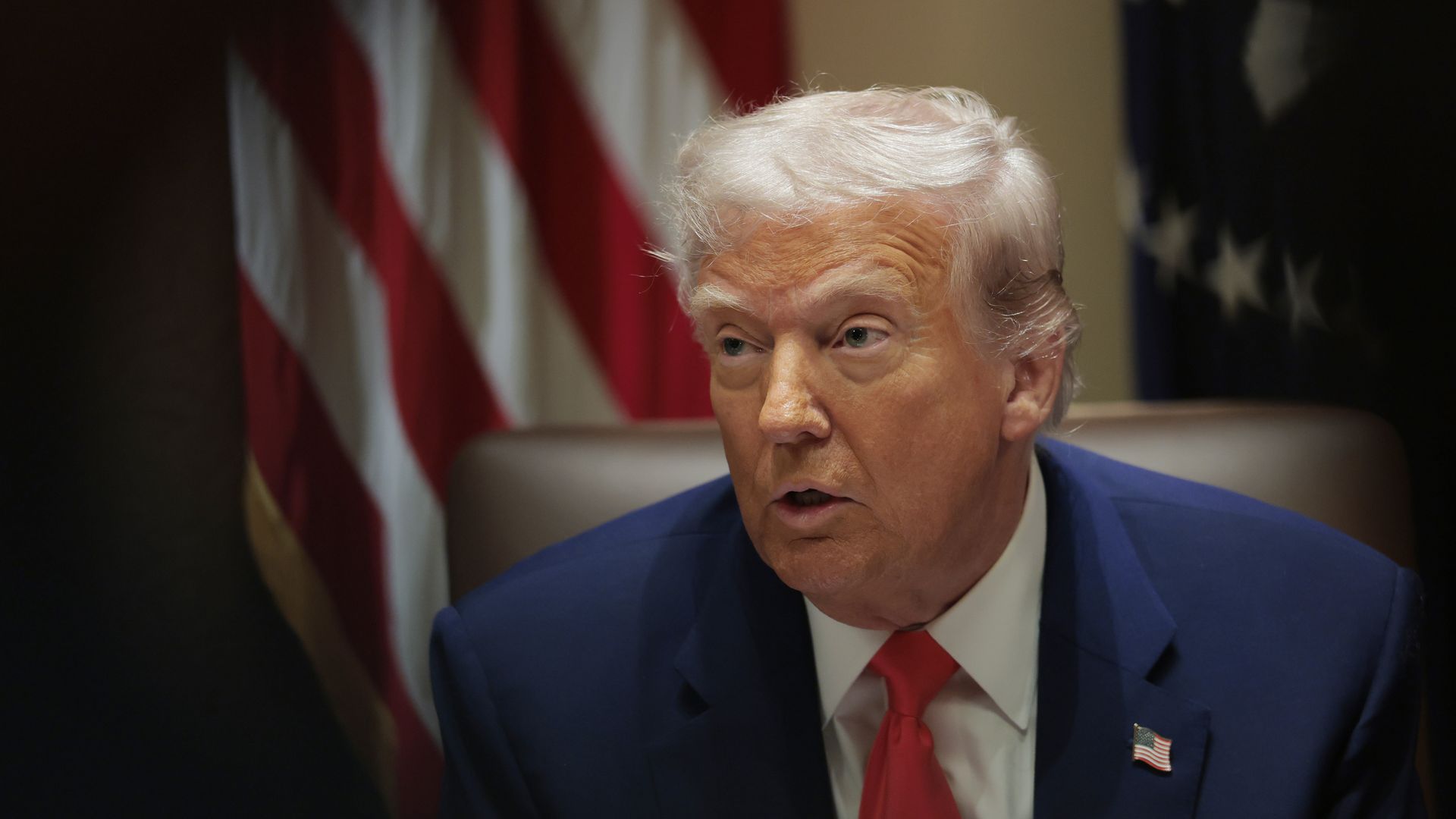 Trump sits at a chair with an American flag behind him 