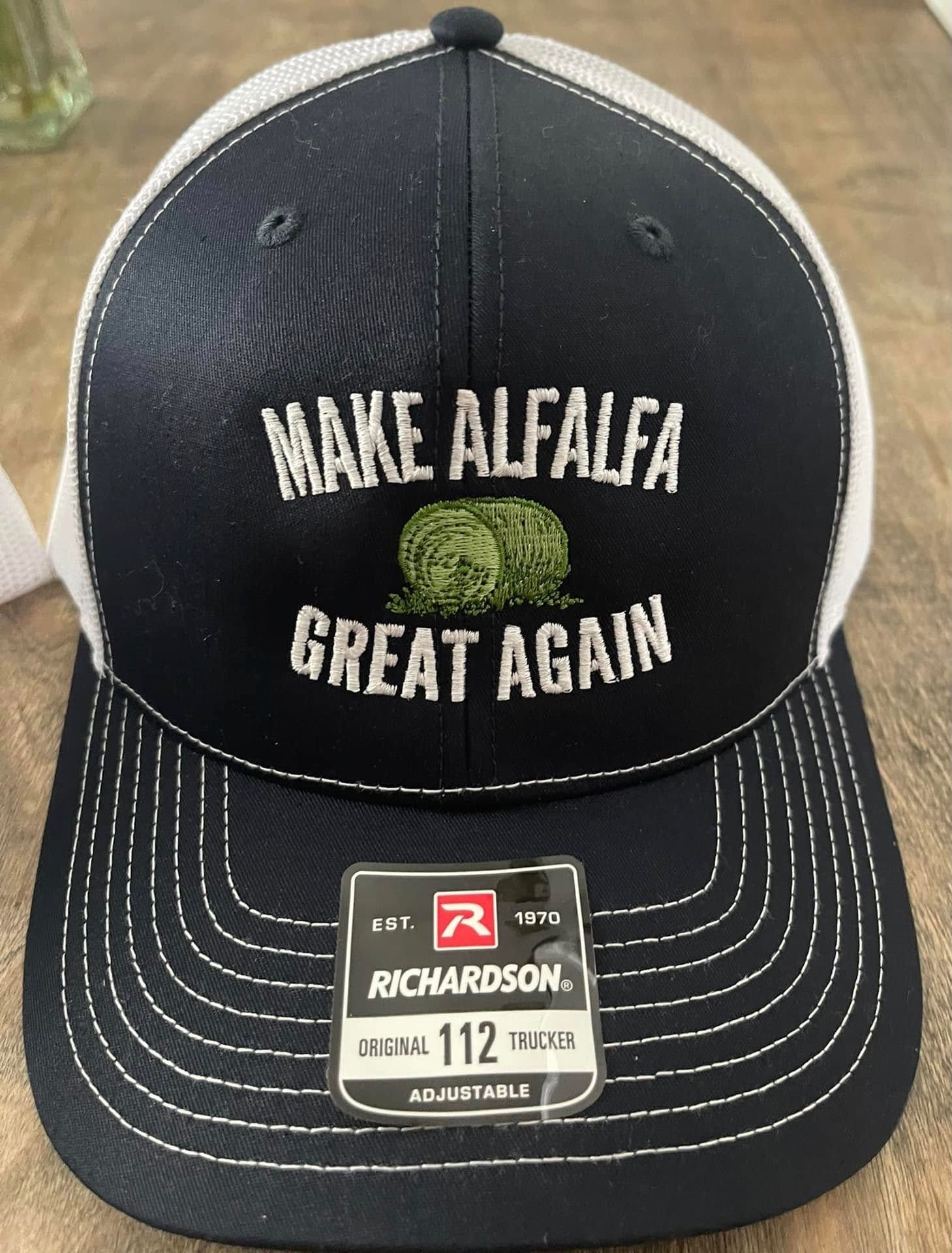 A black trucker hat is embroidered with "MAKE ALFALFA GREAT AGAIN" with an image of a round hay bale. 