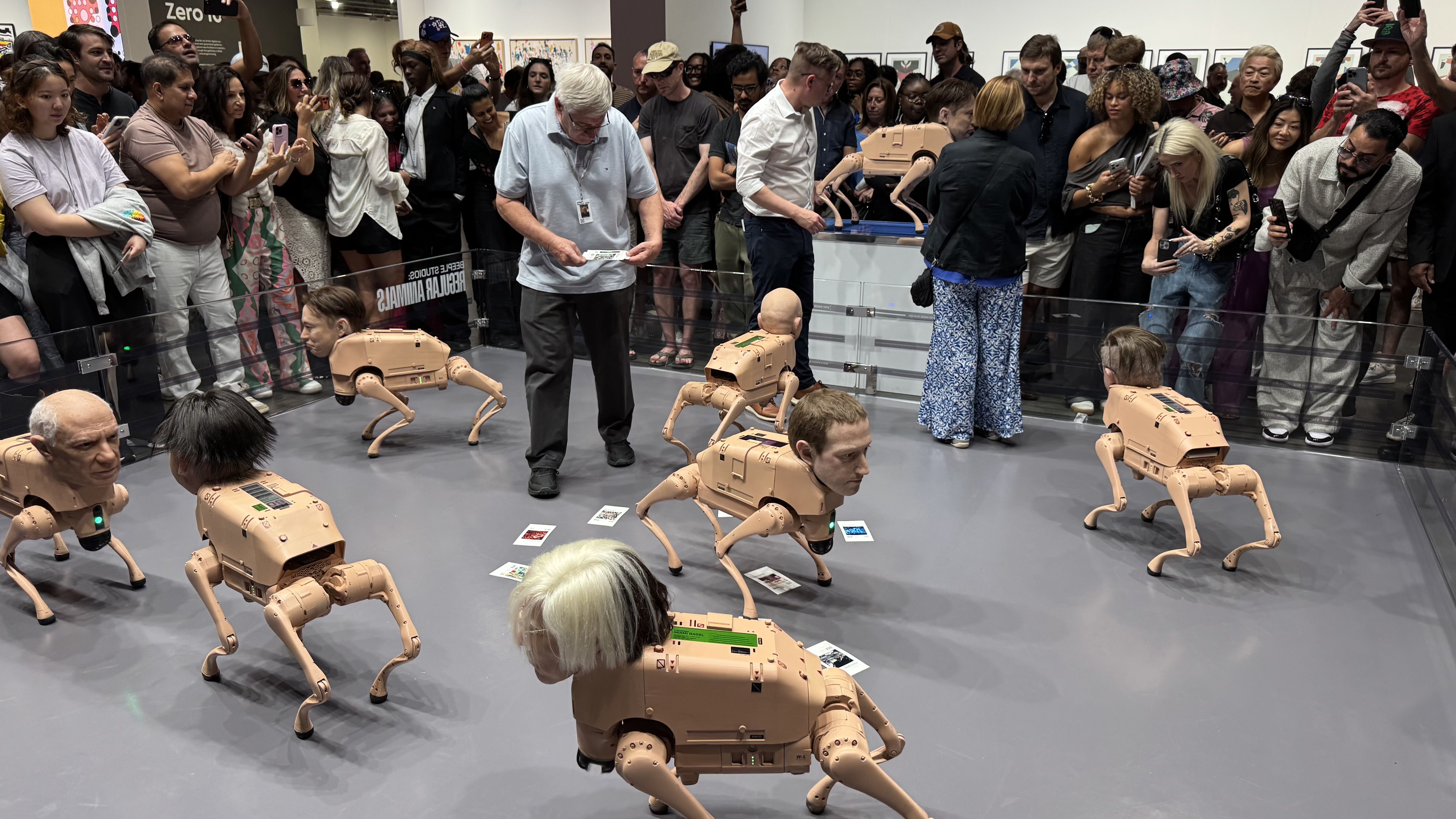 Crowd surrounds exhibit of robots with human heads and dog-like bodies in a gallery, people taking photos and observing the unusual mixed-media art installation.