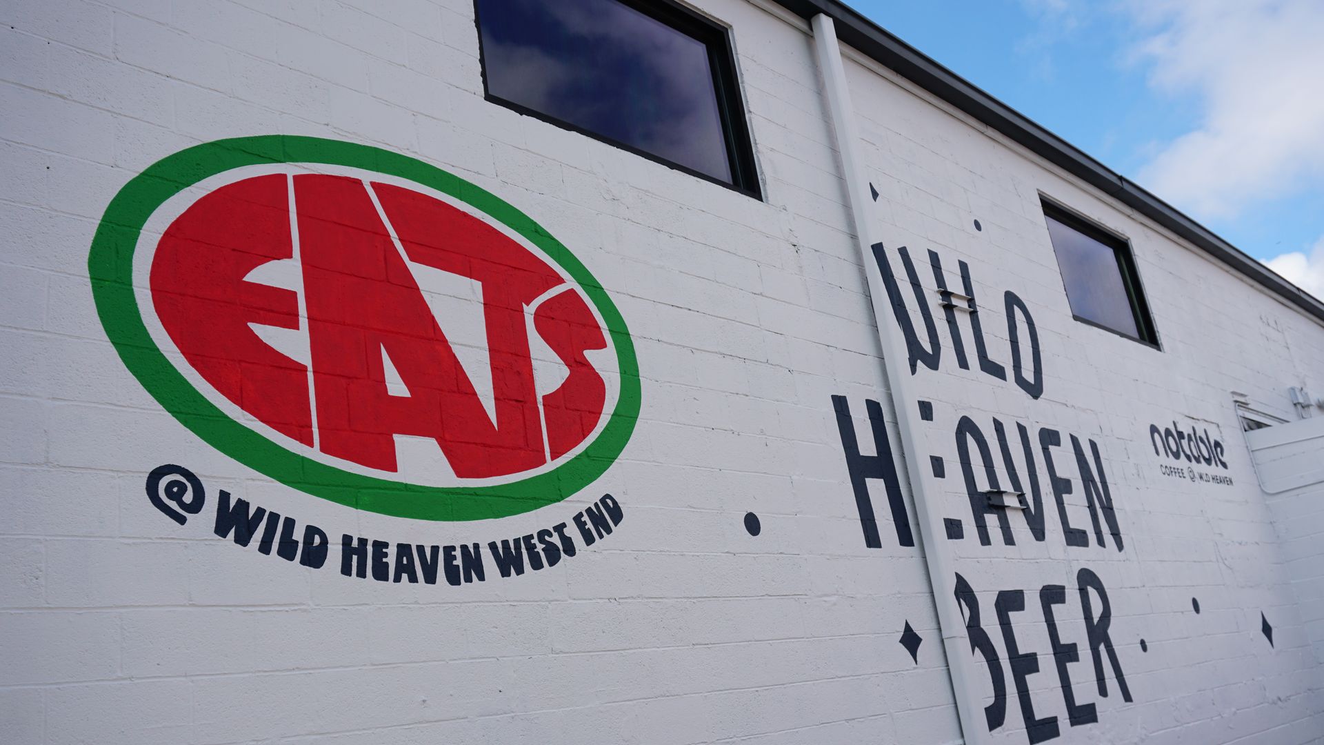 White brick wall with a large circular mural: red center reading EATS in a green ring and text reading "WILD HEAVEN WEST END" to the right of the ring. A windowed building face with blue sky above.