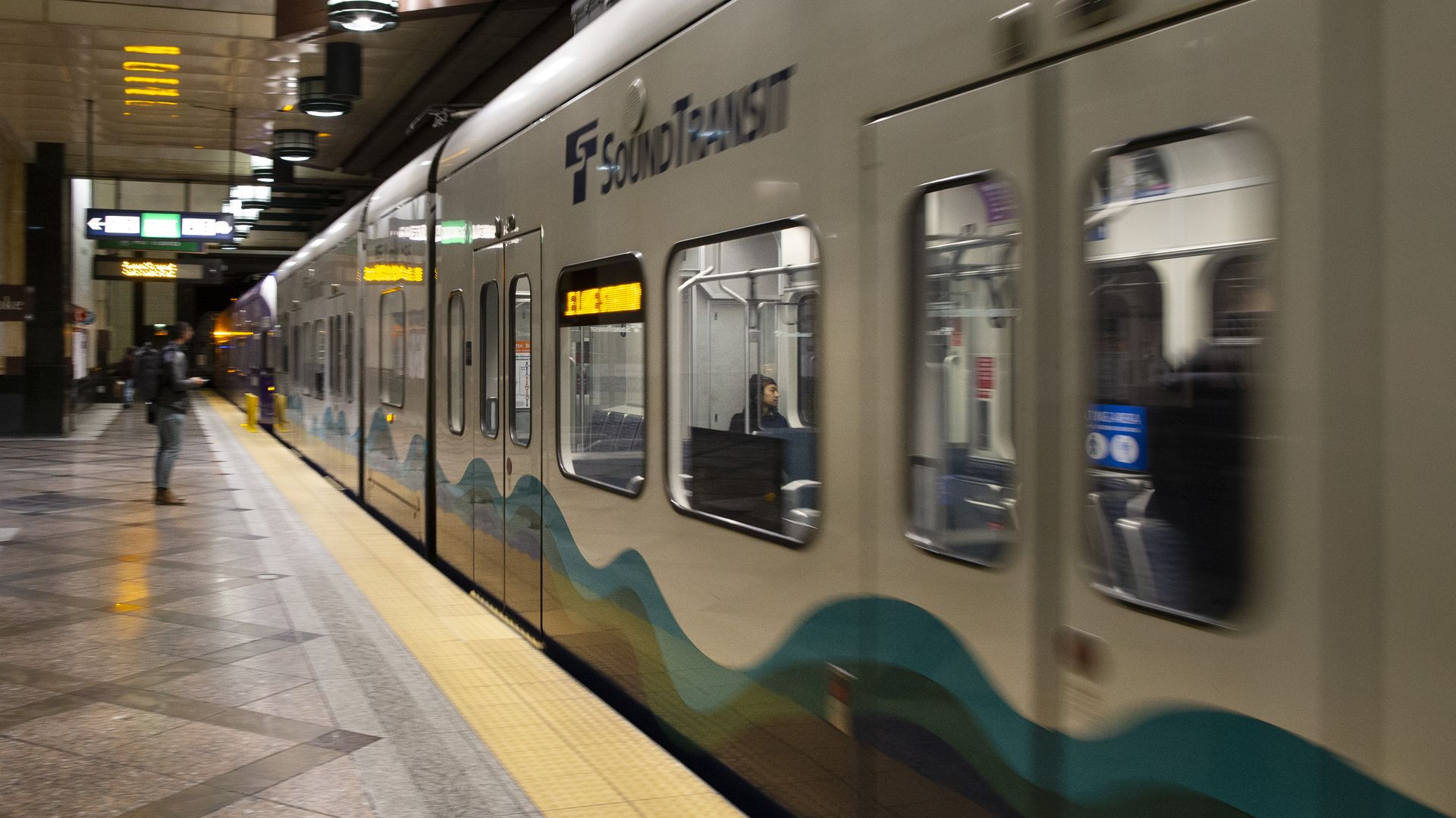 Silver Sounder commuter train with teal and blue wave graphics at an underground platform; a person stands on the left, yellow safety tiles along the edge, with signs overhead.