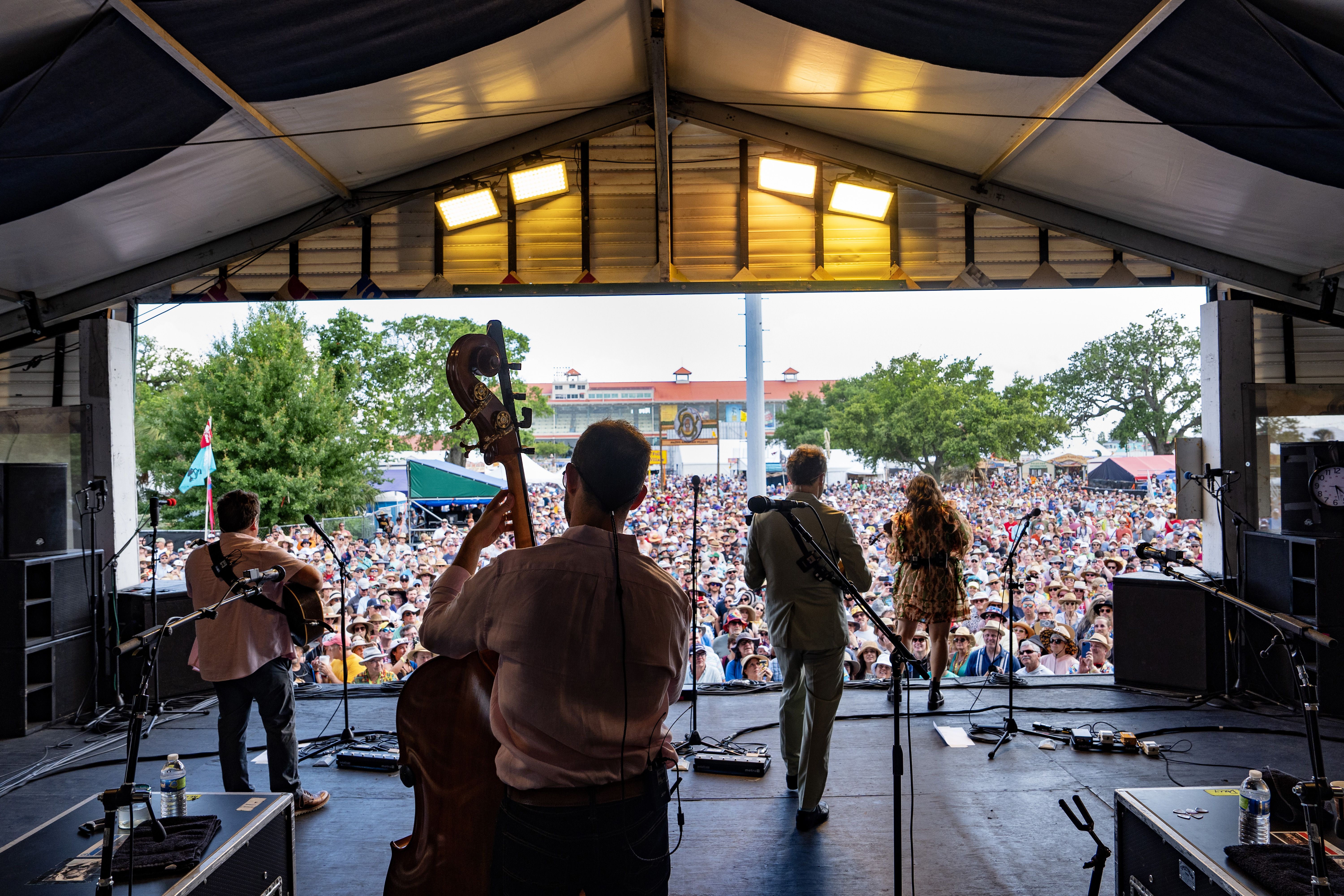 A photo shot from backstage as Nickel Creek performs. The backs of the band-members are seen as they play, and a packed crowd is beyond them.