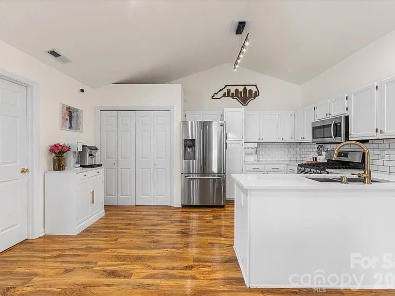 Bright white kitchen with wood flooring, stainless steel refrigerator, white cabinets, subway tile backsplash, and gold faucet. Wall art shows North Carolina with city skyline.