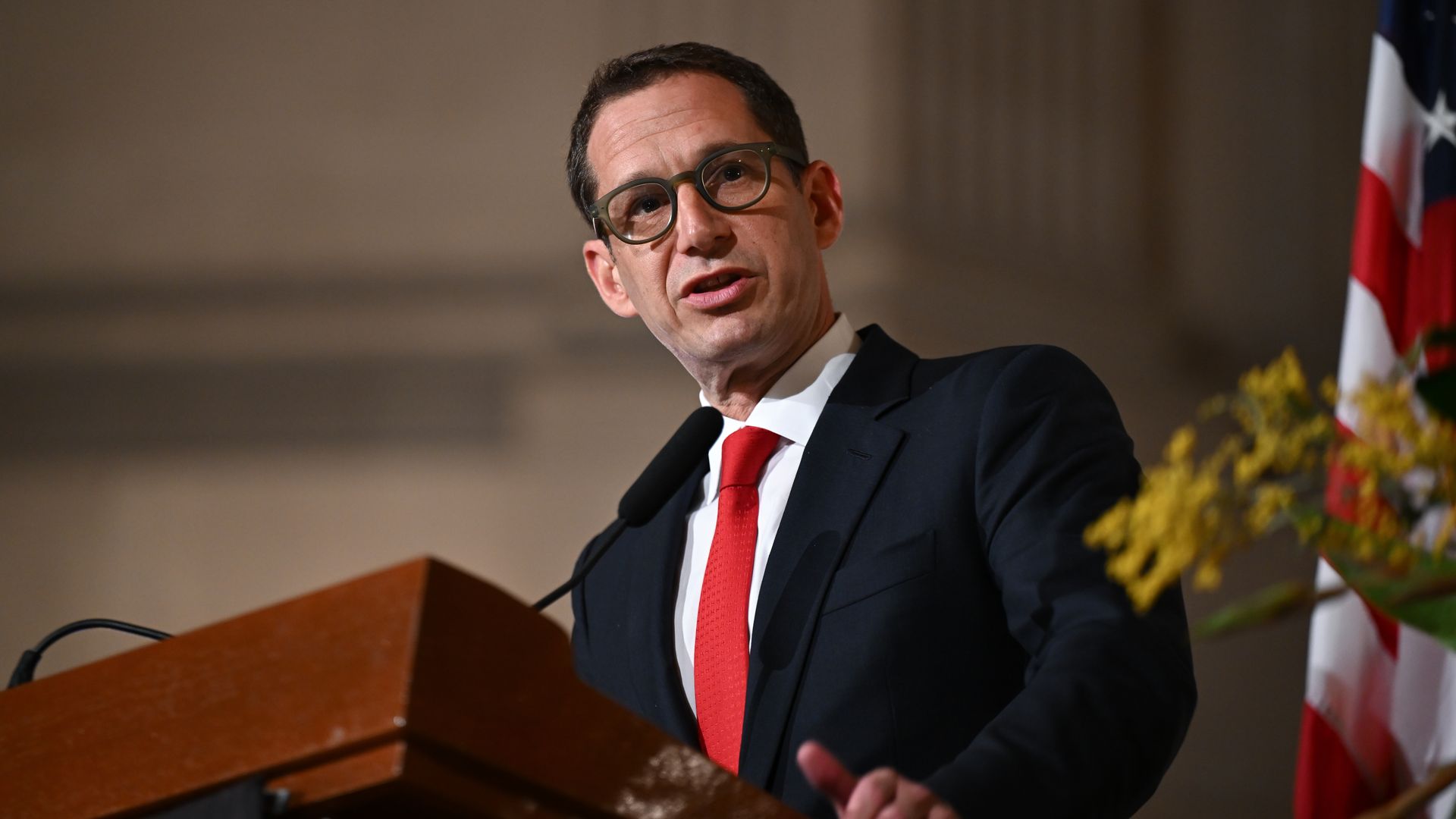 Man in dark suit, white shirt, and red tie speaking at a wooden podium with an American flag and yellow flowers in the background.