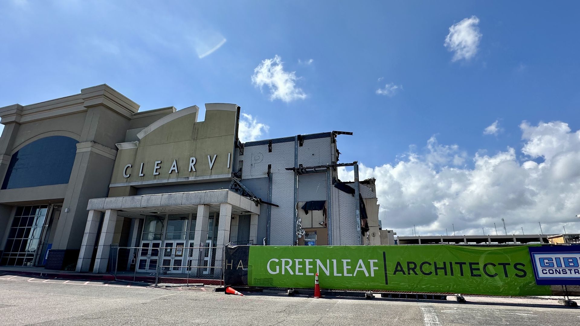 Storefront under renovation with a beige facade and arches on the left, exposed wall and scaffolding on the right, a bright blue sky with scattered clouds, and a green fence banner that reads "GREENLEAF ARCHITECTS".
