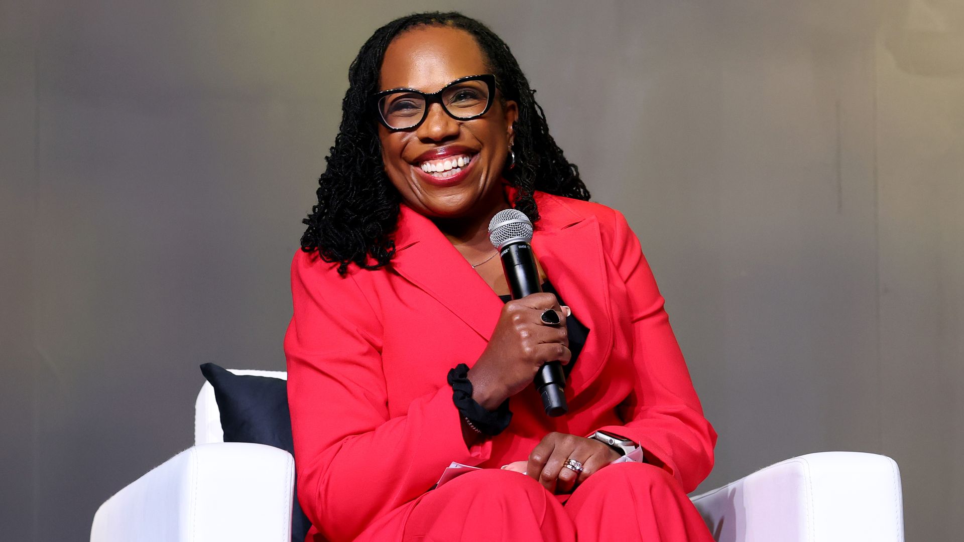 Ketanji Brown Jackson wears  a bright red suit and glasses holding a microphone, sitting in a modern white chair with a black pillow on a gray background.