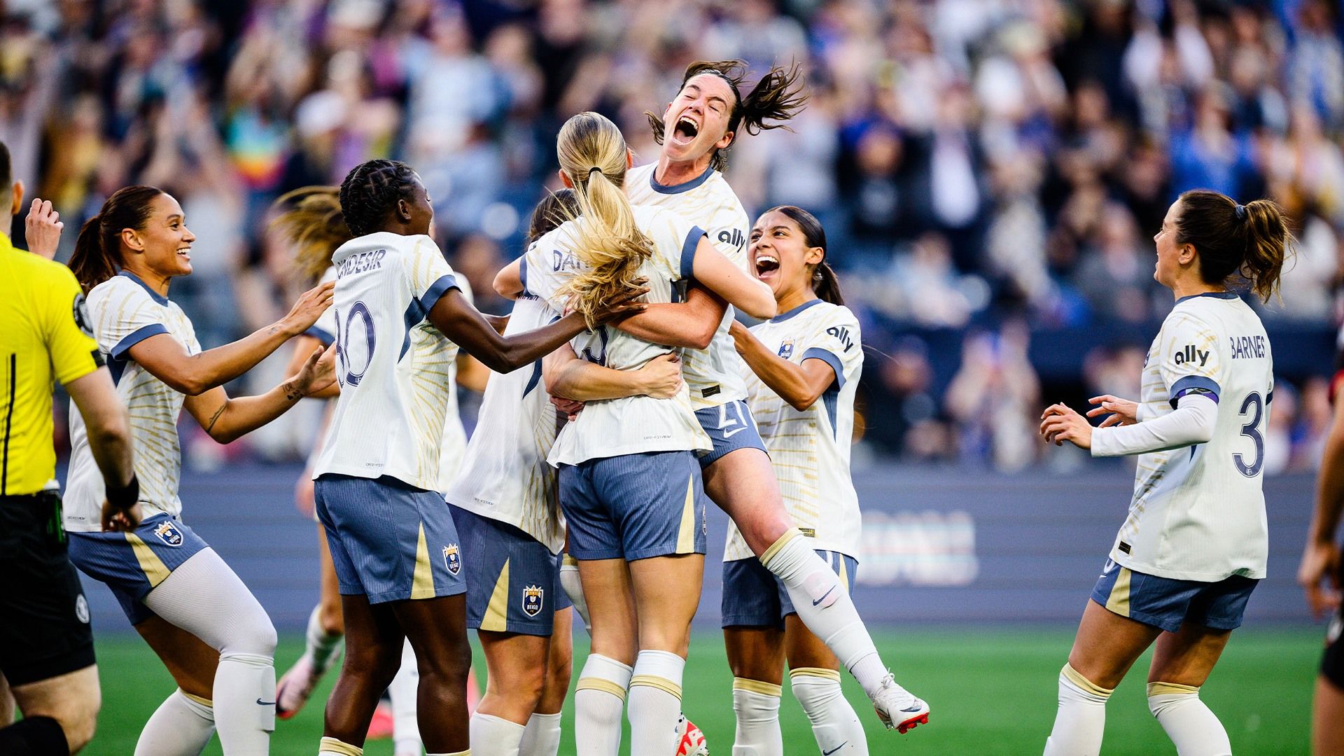 Women's soccer team celebrates together on the field, with players cheering and embracing in front of a lively crowd.