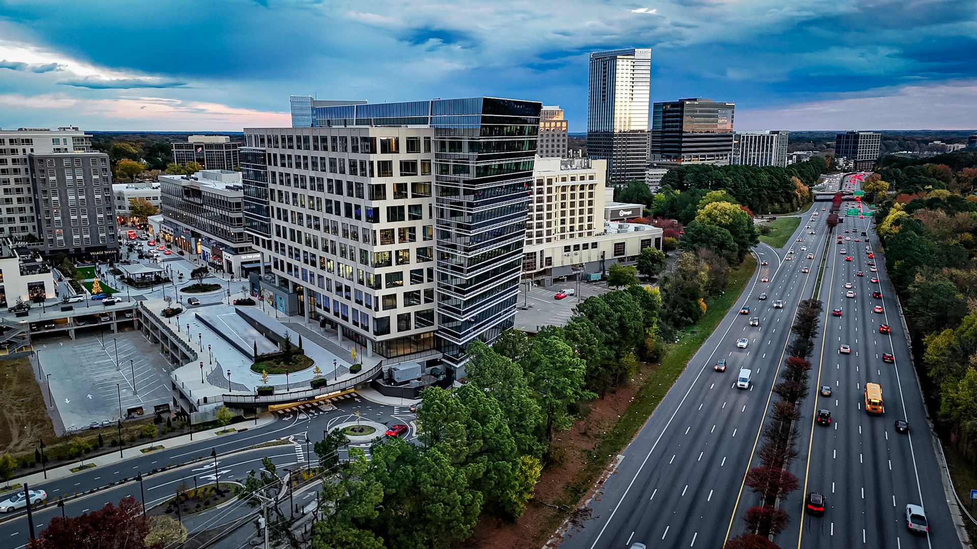 A modern cityscape with office buildings beside a busy multi-lane highway, surrounded by green trees under a blue cloudy sky during dusk.