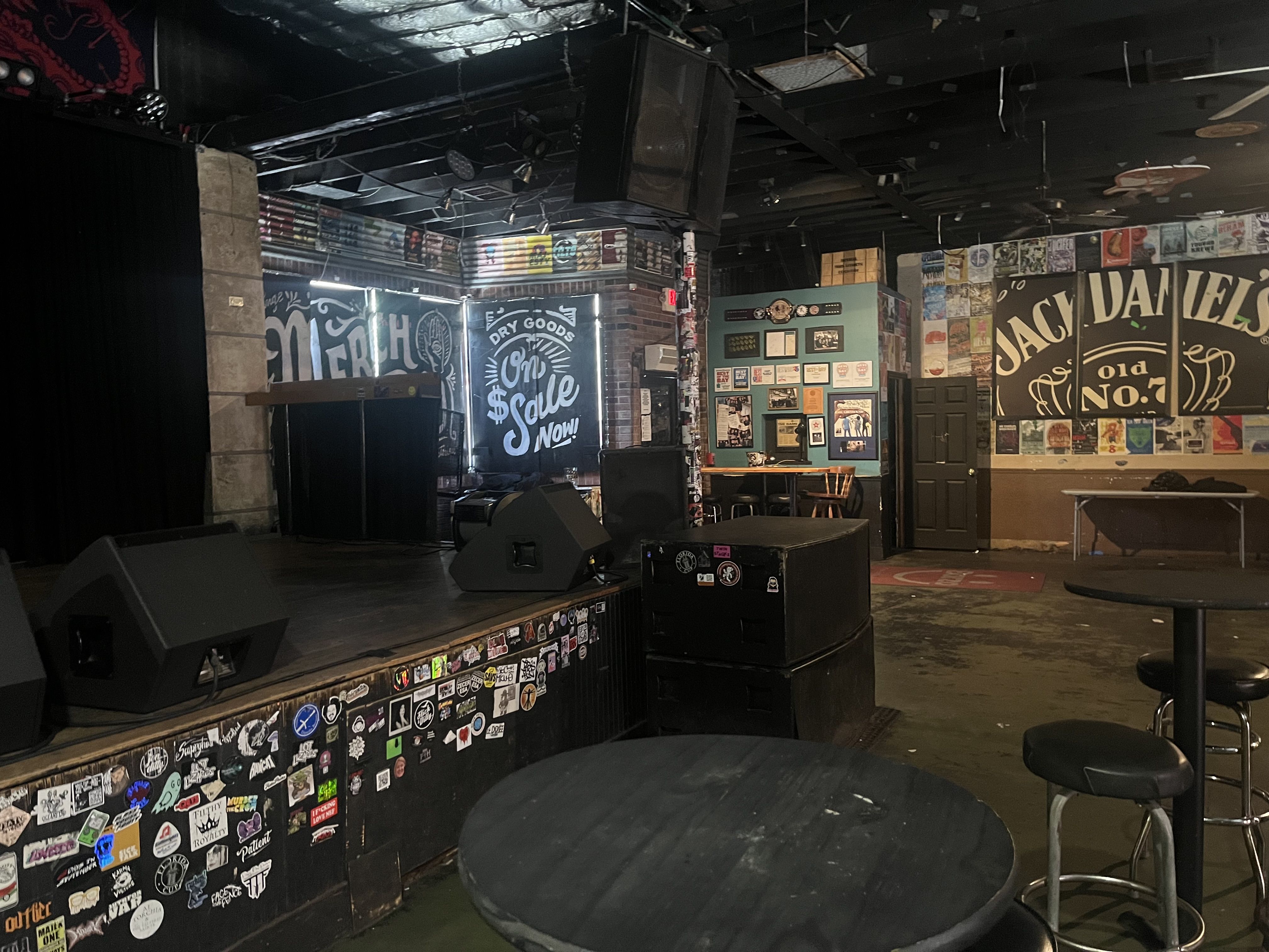 Dim bar stage with black ceiling and exposed beams; left curtain, walls stacked with posters and stickers; right side shows a Jack Daniel's Old No. 7 sign; round tables and stools nearby.