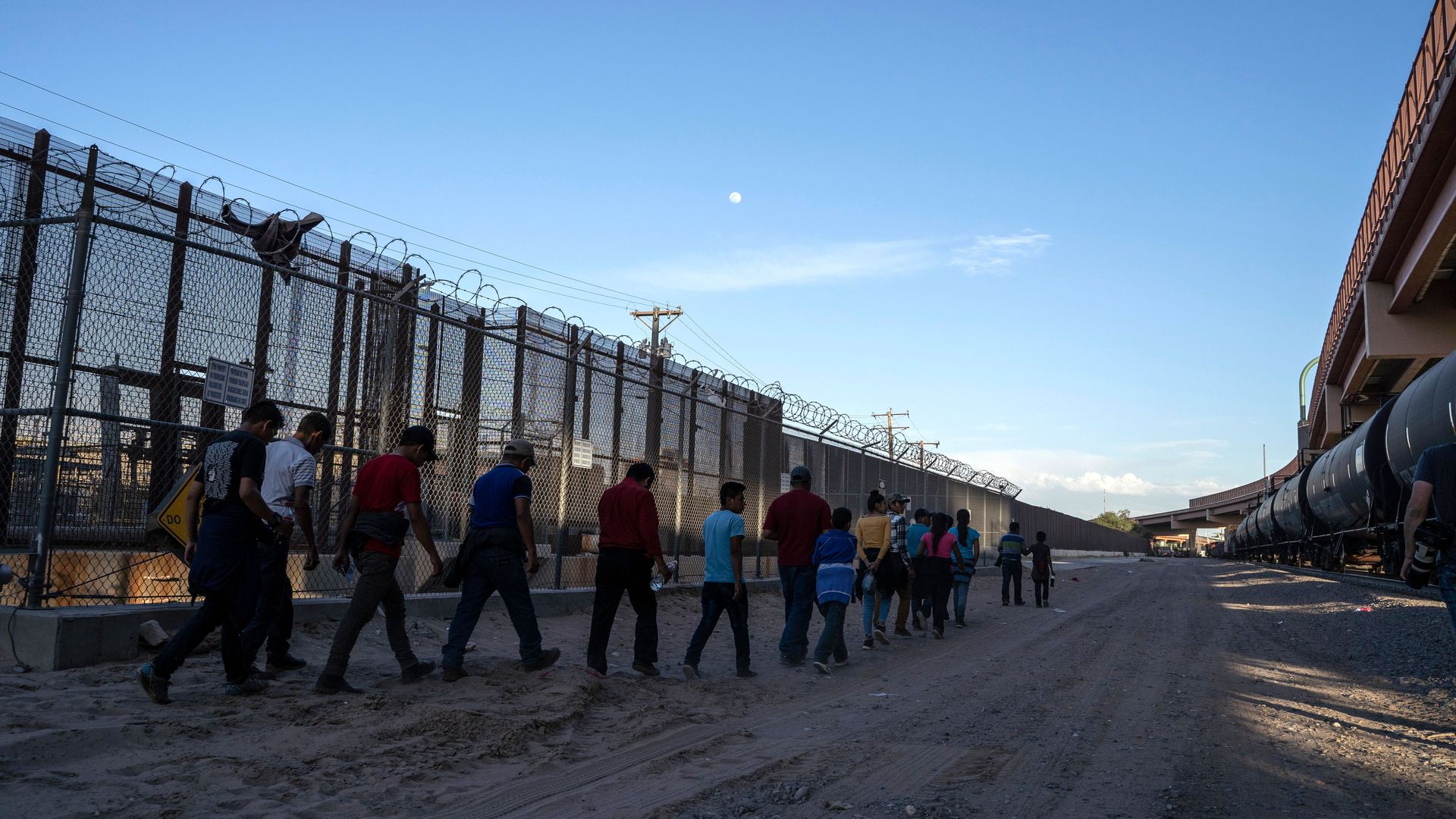 In this image, a line of migrants walk outside near a tall chain link fence.