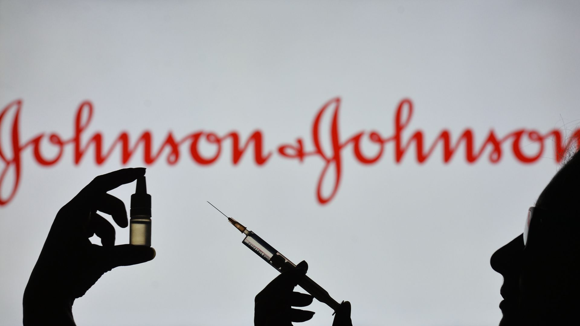 In this photo illustration a silhouette of a man holding a medical syringe and a vial seen displayed in front of the Johnson and Johnson logo on a screen. 