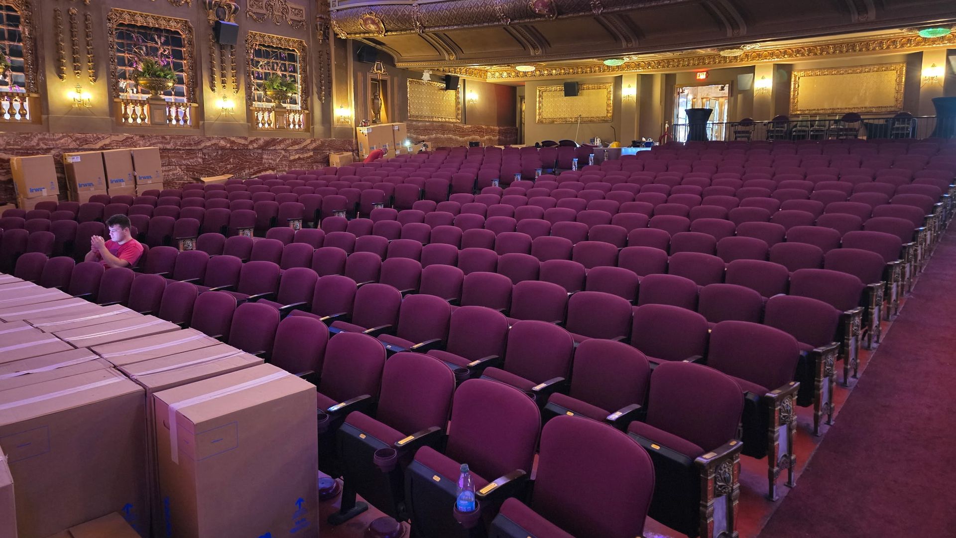 Empty theater with rows of maroon seats, ornate gold and beige walls, and large decorative panels. A few cardboard boxes are stacked on the left, and one person in a pink shirt sits among the seats.